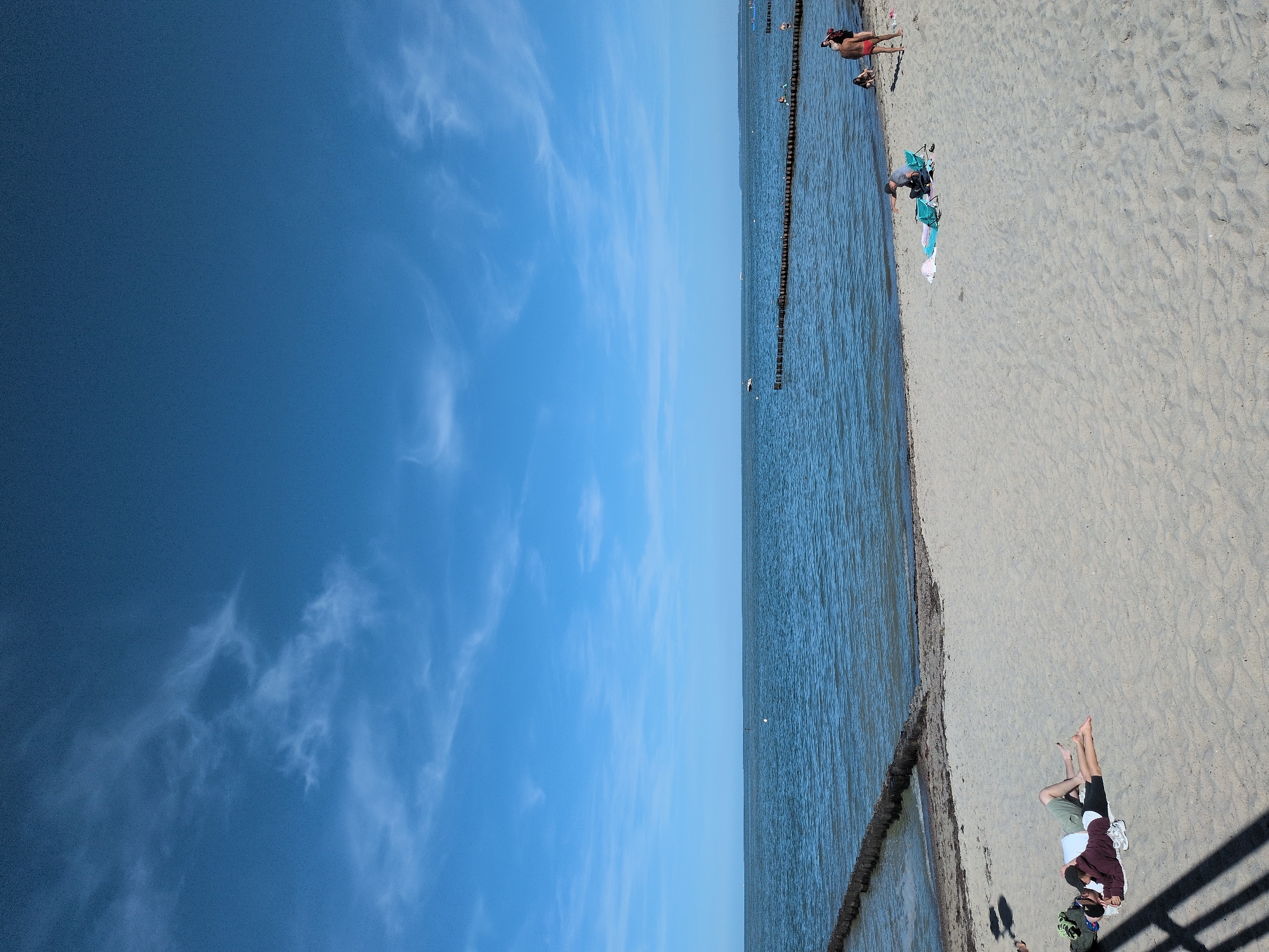 The image depicts a sunny day at the beach in Kühlungsborn, Germany. In the foreground, two individuals are lying on the sand, facing away from the camera, seemingly relaxing. One person is wearing a maroon hoodie and dark shorts, while the other is wearing a white t-shirt and green shorts. To their left, a green bag is placed on the sand. Further down the beach, several other individuals are visible. One person is sitting in a chair by the water's edge, while another, wearing red shorts, stands near them. A few more figures are scattered along the shoreline. The sand is light brown and appears smooth, with some scattered seaweed near the water.
The sea stretches out in the background, a calm blue with gentle waves. A series of wooden breakwaters, arranged in parallel lines, extend from the shore into the water, breaking the monotony of the sea. A small white buoy is visible in the distance, and further out, a boat is sailing. The sky above is a vibrant blue, with wispy white clouds scattered across it, suggesting fair weather. The overall scene is one of a peaceful, leisurely day at the coast.
