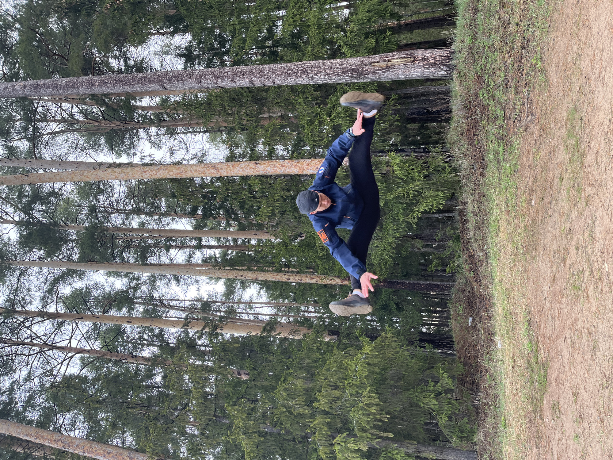 The image, taken in Chaikovsky, Russia, depicts a male individual engaged in a dynamic, mid-air pose within a natural forest environment. The subject is a young man, caught in what appears to be an athletic jump, kick, or acrobatic split, with his left leg extended high to the side and his body angled.
He is dressed in practical athletic attire, including a dark blue zip-up jacket with subtle orange detailing and what appear to be small, indistinct patches or logos on the sleeves and chest. He wears black athletic pants, a dark baseball cap worn backward, and dark-colored sneakers with light soles. His arms are outstretched for balance, with his left hand near his extended left foot and his right arm reaching forward.
The setting is a coniferous forest, characterized by numerous tall, straight tree trunks, likely pine or spruce, that form a dense backdrop. The sky, visible between the tree canopies, is bright and overcast, indicating daytime with soft, diffused lighting. To the right of the person, there's a gentle incline of ground, covered with sparse green grass and some dry undergrowth, leading to a light brown dirt path or road. The overall scene suggests an active moment in a rugged, outdoor landscape. There is no discernible text visible on clothing or in the surroundings.
