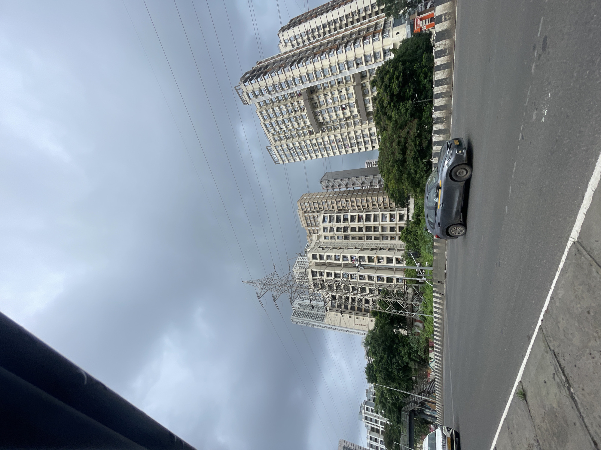 An overcast, gloomy sky looms above a cityscape in Mumbai, India. In the foreground, a multi-lane asphalt road stretches horizontally, with a gray car in the right lane and a white car in the left lane. The road is bordered by a concrete divider with black and white stripes. Behind the divider, lush green trees form a dense canopy. Tall, modern apartment buildings, characterized by their numerous windows and balconies, rise behind the trees. A prominent electricity pylon stands tall amongst the buildings, with power lines crisscrossing the sky. The architecture of the buildings suggests a contemporary urban environment.