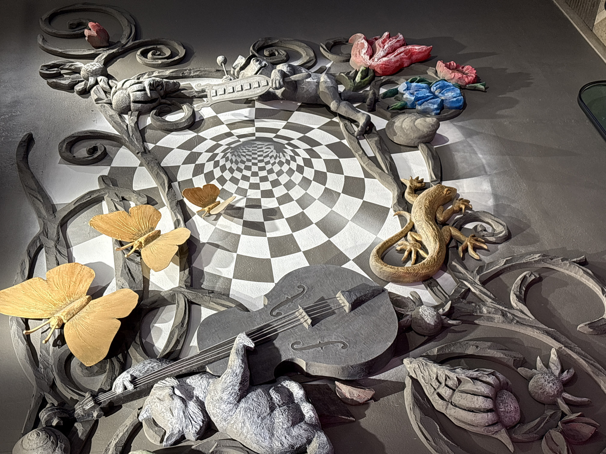 The image depicts a three-dimensional art installation mounted on a wall, possibly within an interior space. The installation has a dreamlike, surreal quality, with various sculpted elements arranged within a frame.
The background is a checkered pattern that appears to recede into a tunnel-like vortex. Above the checkerboard are sculpted figures of a man and a variety of flowers. Below the checkerboard pattern are a stringed instrument that appears to be a cello, a sculptural figure, butterflies, a lizard, and various floral designs.
The installation has elements reminiscent of the artwork of Salvador Dali and is located in Yekaterinburg, Russia.