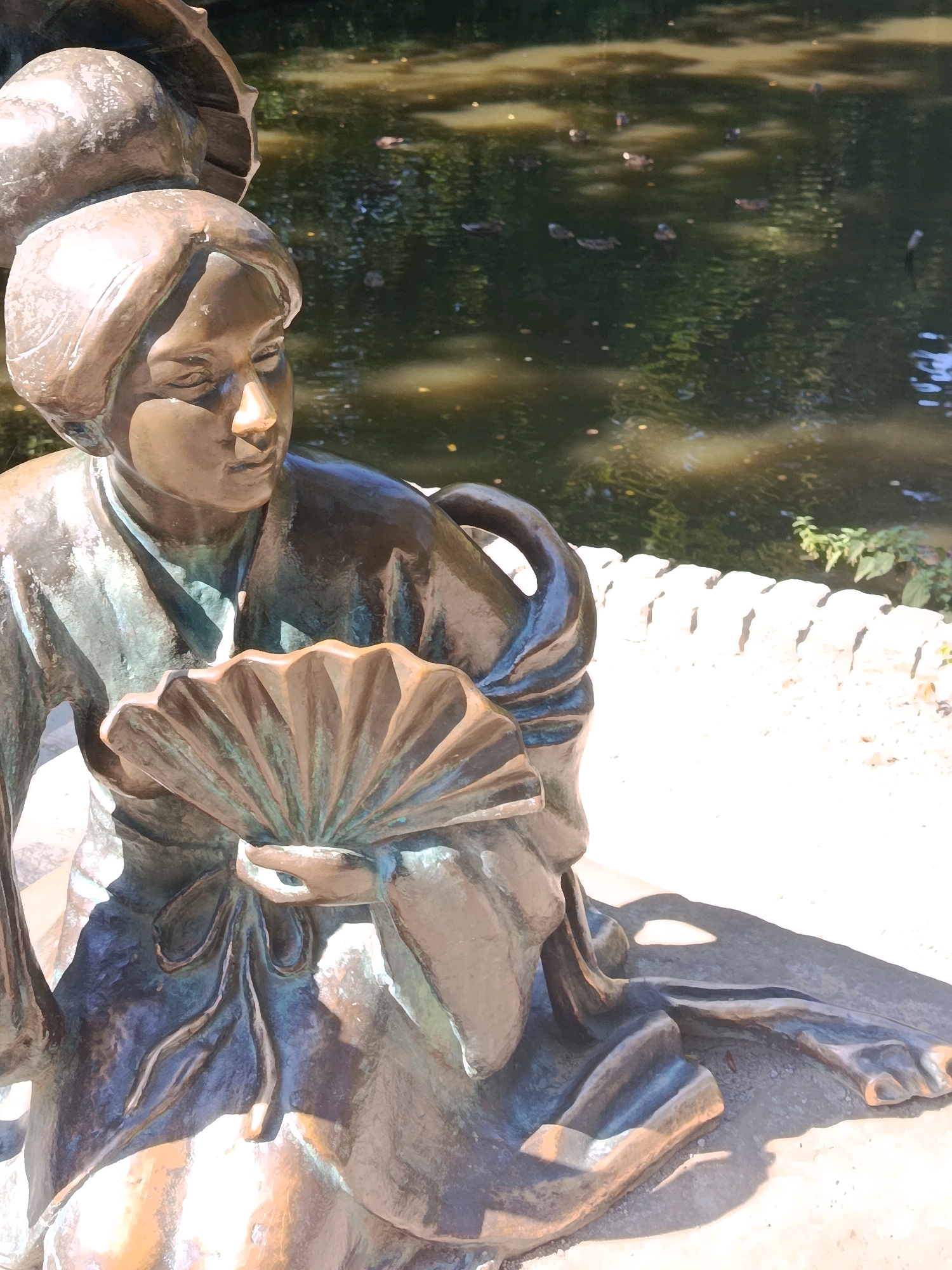 This image shows a close-up of a bronze statue of a young woman holding a fan. The statue is in a seated position, with her legs bent and crossed. She is wearing traditional clothing, with flowing sleeves and a sash. The statue is made of weathered bronze, with a patina that gives it a greenish-blue hue in some areas. The woman's face is serene, with her eyes downcast. She is looking at the fan, which is open and held in her hands. The fan is intricately detailed, with delicate folds.
The statue is situated outdoors, next to a body of water that appears to be a pond or a small lake. The water is dark and reflective, with ripples on its surface. Several ducks are visible swimming in the water. Behind the statue, a low, white stone wall can be seen, possibly marking the edge of the pond or a garden. The background is filled with lush green foliage, suggesting a park or garden setting. Sunlight is casting shadows on the statue and the ground, indicating it is daytime.
The statue is located in the Arboretum "Oleksandriya" in Bila Tserkva, Ukraine. This is a well-known botanical garden and park, which features numerous sculptures and decorative elements. The statue of the woman with the fan is one of its many attractions.