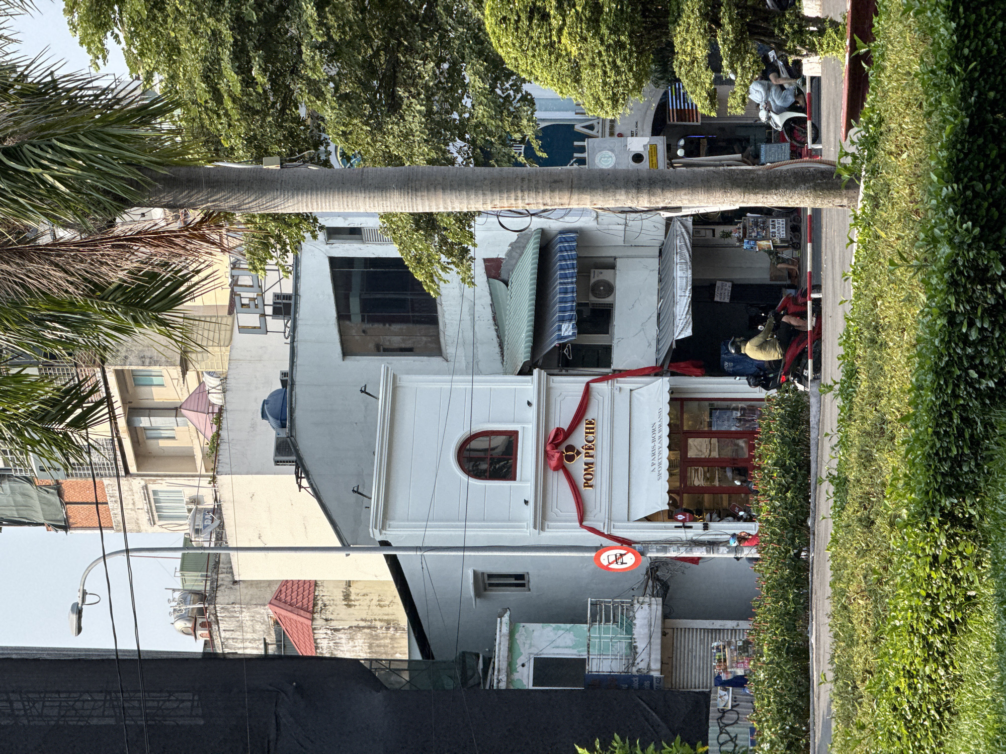 The image shows a street scene in Thủ Đức, Vietnam. The main focus is a white building with a sign that reads "Pom Pêche," adorned with a red ribbon. A round window is located above the sign. To the left of the building, there is a taller structure with windows and a light fixture. A palm tree is positioned partially in front of the white building, its trunk and fronds are visible. In front of the building and partially obscured by the green hedge is a street with motorbikes parked. A man is sitting on one of the motorbikes. The street is bordered by a dense green hedge. Other buildings are visible in the background, along with utility poles and wires.
