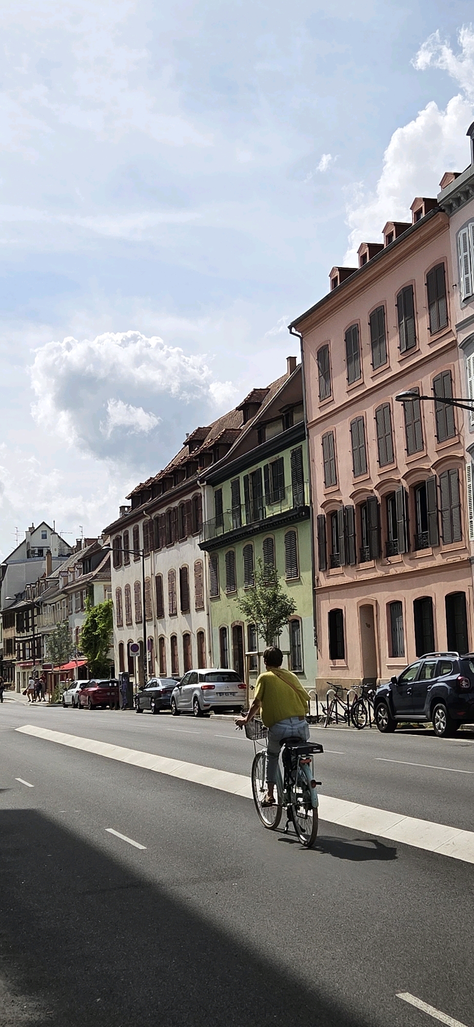 In Strasbourg, France, a person is riding a bicycle on a street lined with colorful buildings. The cyclist, wearing a yellow shirt and blue jeans, is in the foreground, moving away from the camera. The street has a white lane divider and a white raised section on the right side. The buildings on the left and right sides of the street are of various colors, including pink, green, and white, and feature traditional architecture with shutters on the windows. Several parked cars are visible along the street, and trees are planted in front of some buildings. The sky is partly cloudy with fluffy white clouds.