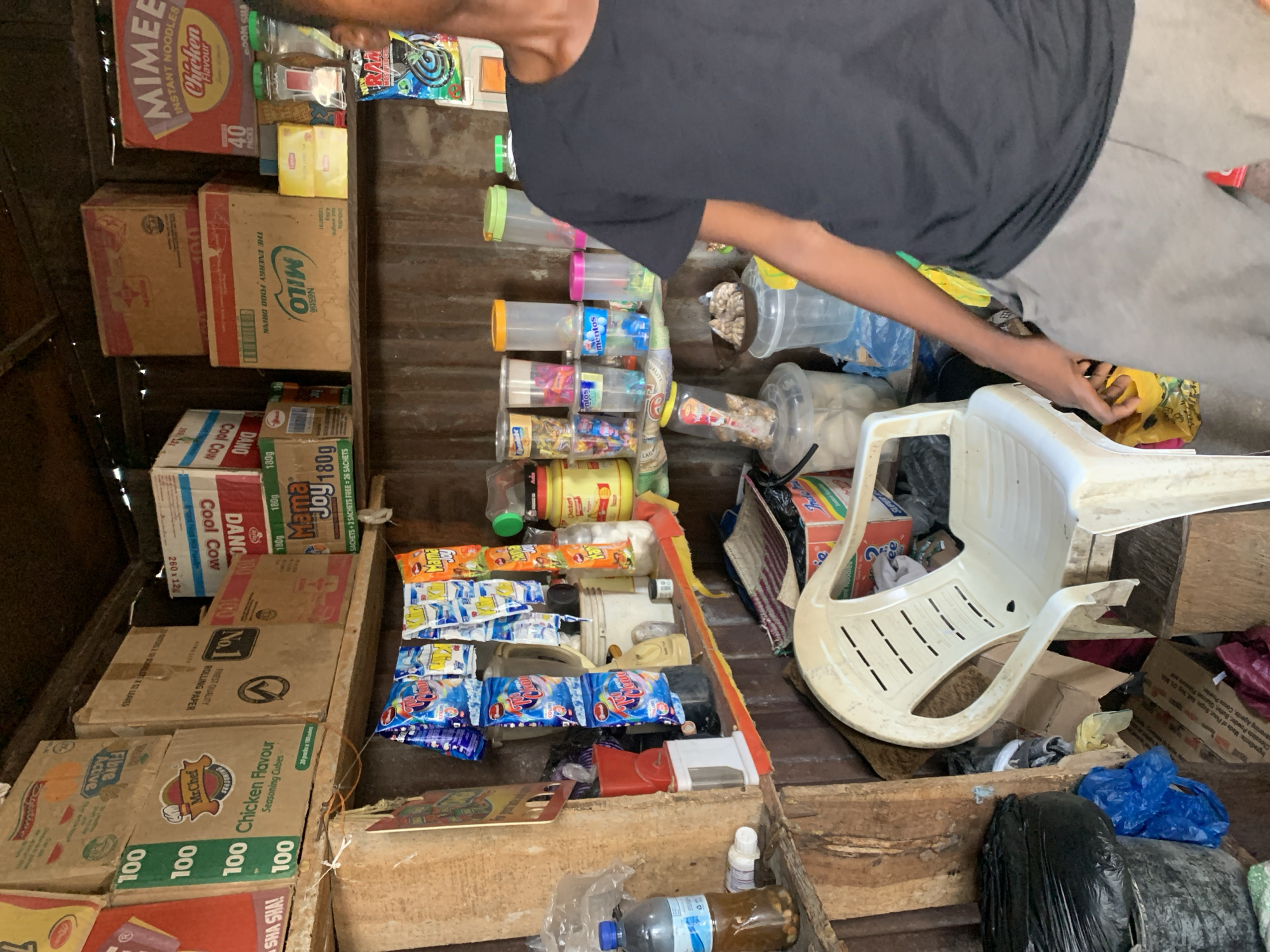 A cluttered interior in Adankolo, Nigeria. An individual wearing a dark gray T-shirt is partially visible, reaching toward a white plastic chair. The space is filled with various food products, including boxes of Milo, Mimee Instant Noodles, Cool Cow, Dano, and Mr. Chef Chicken Flavour seasoning cubes. Numerous small packets and plastic containers filled with goods are present. The environment appears to be a storage or sales area within a structure with corrugated metal walls.