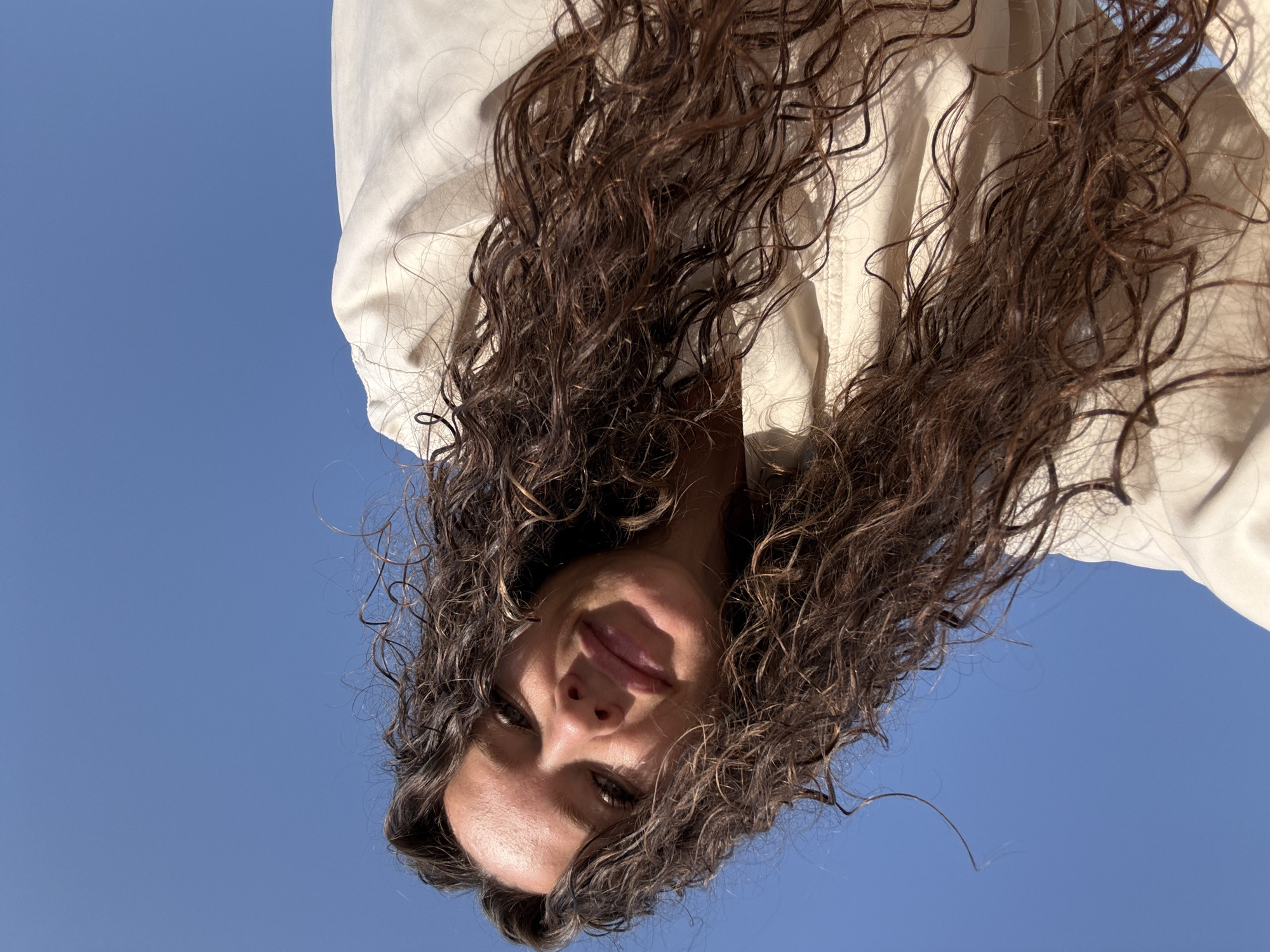 An outdoor, low-angle, waist-up selfie shows a woman with long, dark, curly hair against a bright blue sky. She is wearing a light-colored collared jacket. The woman is looking at the camera and smiling slightly. Her face is illuminated by sunlight, casting shadows on her skin. The background is a clear blue sky, suggesting a sunny day. There is no indication of the city of Dubai or the United Arab Emirates.