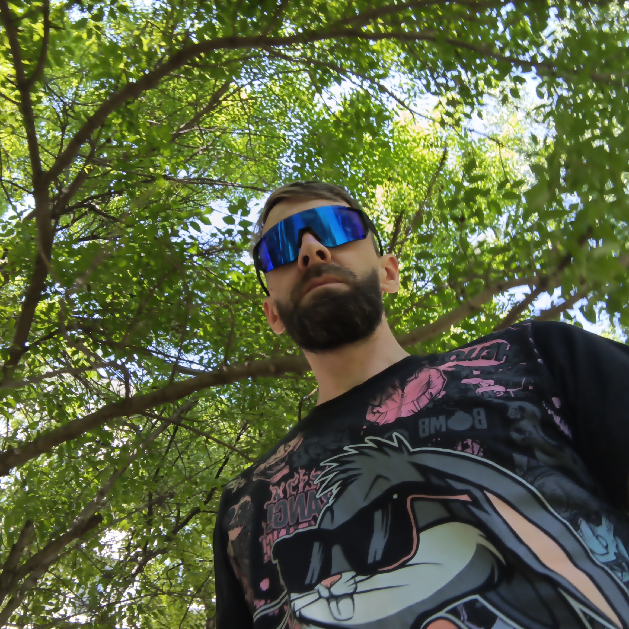 A man with a beard and reflective blue sunglasses is photographed from a low angle, looking down at the camera. He is wearing a black t-shirt with a cartoon design featuring a rabbit wearing sunglasses. The background consists of dense green foliage and tree branches, with glimpses of the sky visible through the leaves. The photograph was taken in the city of Chaikovsky, Russia.