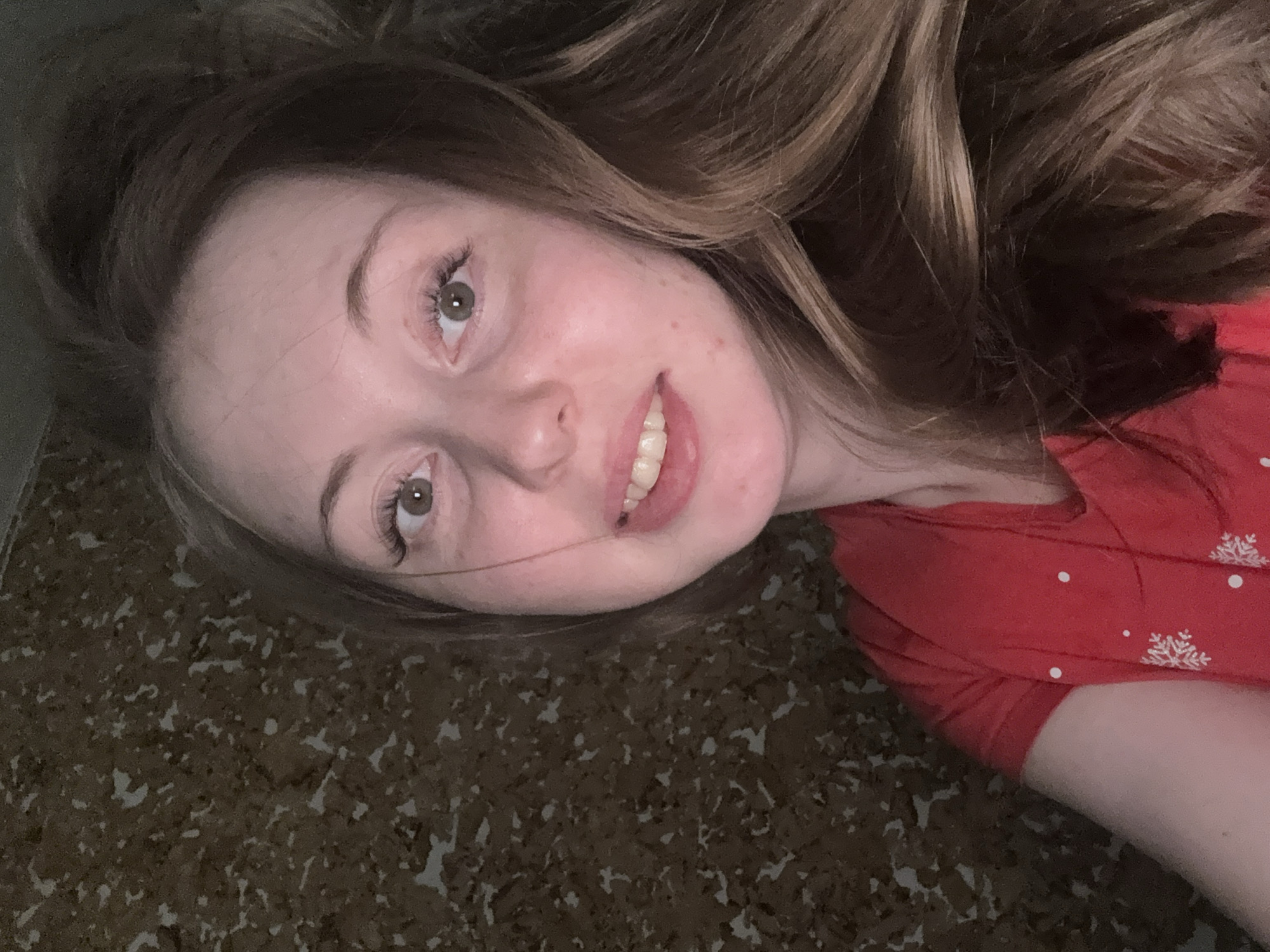 A young woman with light brown hair and light skin is lying on a patterned surface, possibly a rug or floor covering. She is wearing a red top with white snowflake and polka dot patterns. She is smiling and looking at the camera. The location is Unknown, Unknown.