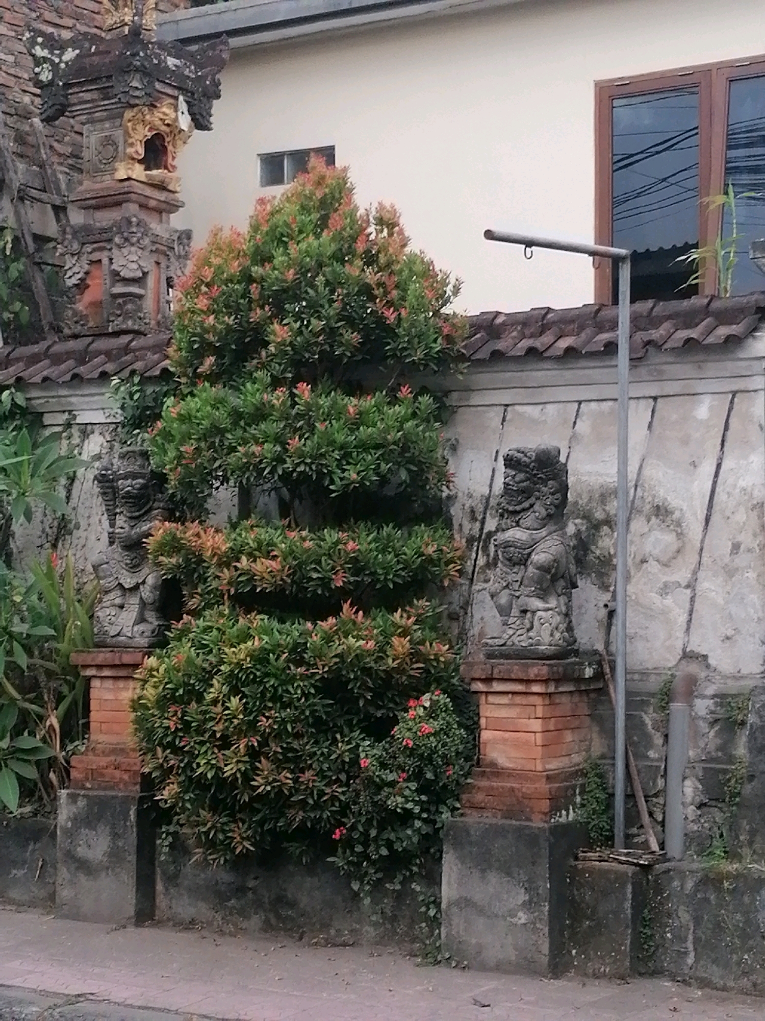 This image shows a decorative stone statue, possibly a guardian figure, situated in front of a textured wall and a lush green shrub. The statue is placed on a brick pedestal. Behind the shrub, a section of a building is visible, featuring a window and a tiled roof. A metal pole extends upwards from the ground near the wall, with a hook at its end. The overall setting suggests a cultural or historical location in the Ubud District, Indonesia, with elements of traditional architecture and landscaping.