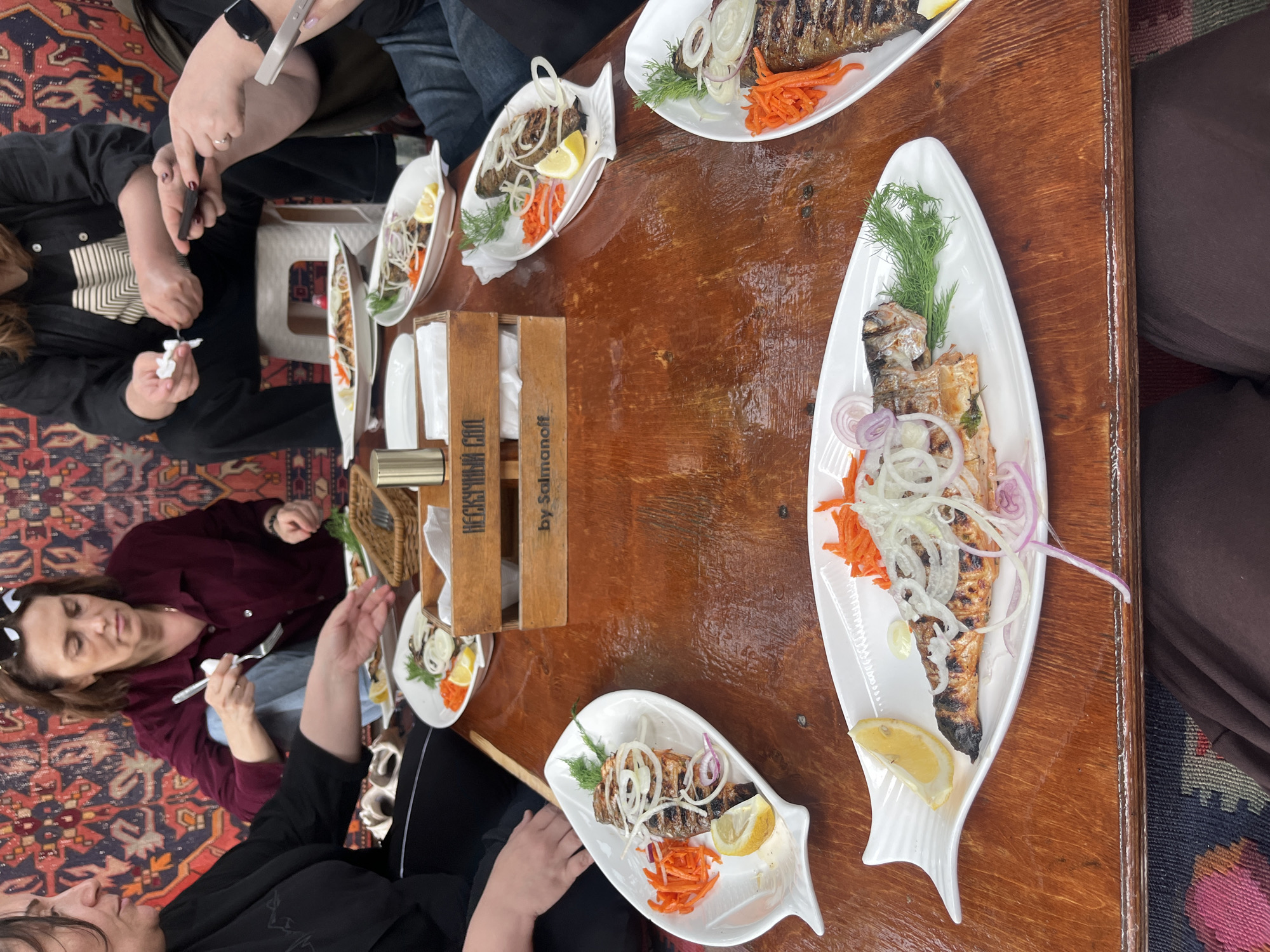 A group of people are seated around a wooden table laden with plates of grilled fish in what appears to be a restaurant or dining setting. The fish dishes are served on white, fish-shaped plates and garnished with lemon slices, shredded carrots, dill, and onion rings. A wooden box with the words "HECKYPHHый COA" and "by Salmanoff" in Cyrillic text is placed centrally on the table. People are interacting, with some holding forks and others with napkins or phones in their hands. The setting is inside, with an ornate carpet visible under the table, suggesting a location that may be in Miatli, Russia, based on the Cyrillic text.