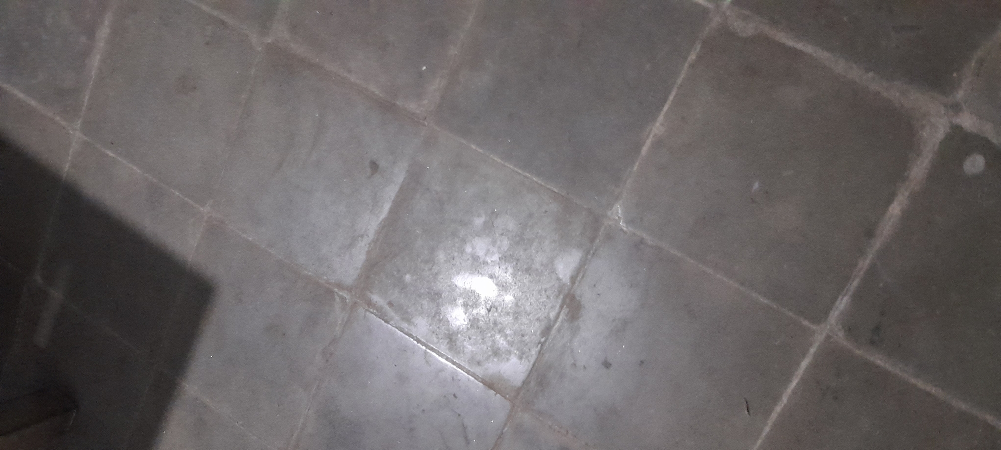 The image shows a tiled floor in what appears to be an indoor environment. The tiles are large, square, and dark in color, with visible grout lines forming a grid pattern. There are some lighter colored smudges or stains on a few of the tiles. In the upper left portion of the image, a dark shadow of an object is cast diagonally across the floor. The lighting suggests the image was taken indoors, possibly in low light conditions. There are no people or specific objects visible that would indicate a particular activity or location within Singkalanyar, Indonesia.