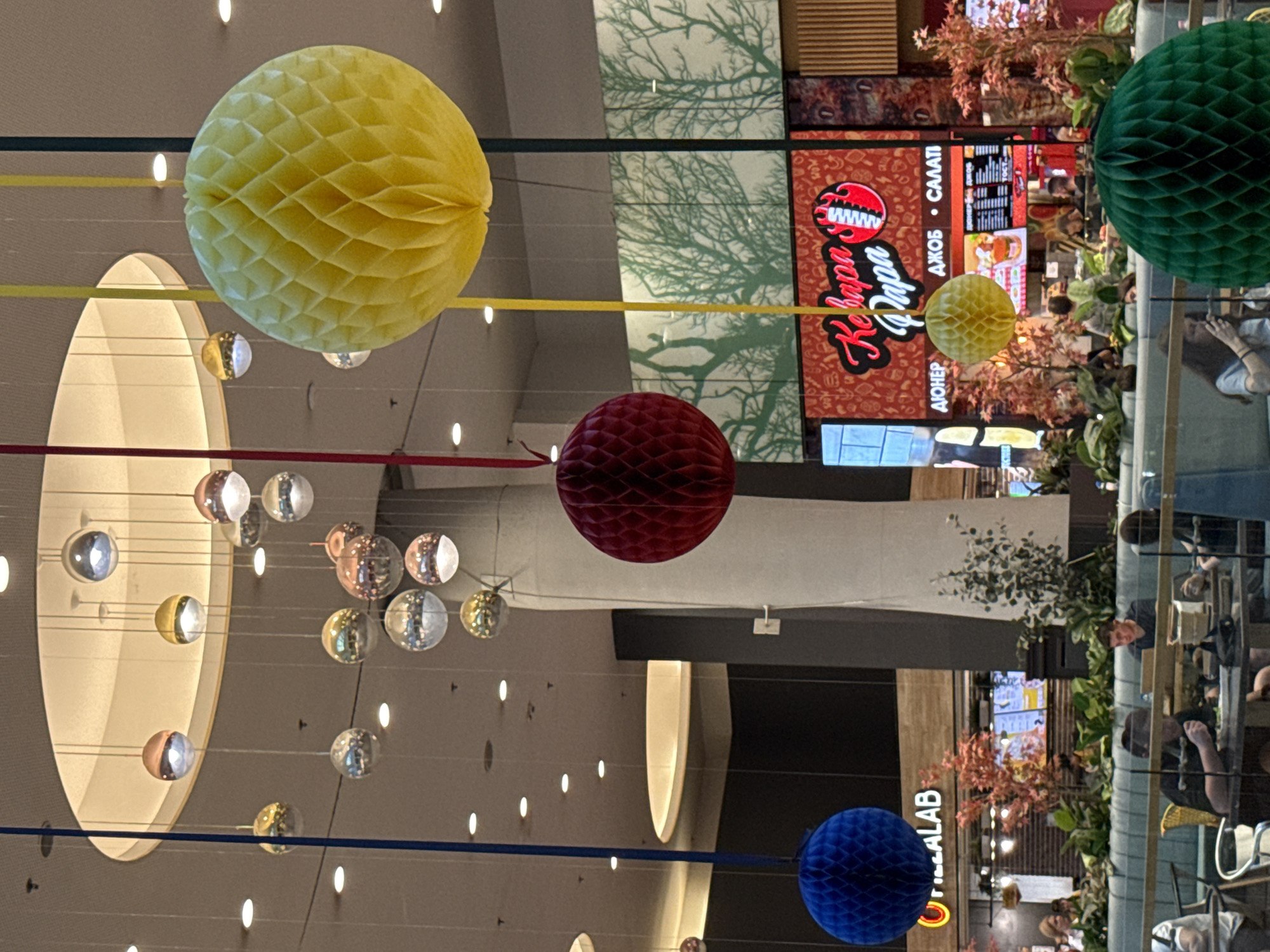 An indoor food court or shopping mall atrium features a high ceiling with recessed circular lighting and a large, light-colored, circular recessed bulkhead. Numerous spherical, reflective light fixtures in silver, gold, and rose gold hues hang at varying lengths from thin wires. Interspersed among these are large, vibrantly colored honeycomb paper balls: a blue one on the far left, a dark red one in the center-left, a large yellow one on the right, and a smaller yellow one in the mid-ground. A green honeycomb ball is partially visible in the bottom right foreground.
The mid-ground features a long, light blue/teal upholstered booth with dark wooden tables. Approximately ten individuals are visible, primarily seated at these tables, engaging in activities such as eating from plates, operating laptops, and looking at mobile phones. Artificial plants and trees with reddish foliage are placed within the seating area and between the food stalls.
In the background, a large mural depicts green stylized tree silhouettes on a white background. Below this, a prominent red illuminated sign for "Кебаба Папа" (Kebapa Papa) features a stylized flaming kebab logo. Beneath the main title, smaller Cyrillic text reads "АЮНЕР • ДЖОБ • САЛАТИ" (Shawarma • Pocket • Salads). To the far left, a partial sign reads "O PIZZALAB". Other food stalls with digital menu screens and wooden paneling line the back wall. A tall, light-colored, possibly fabric-draped column stands on the right, and a dark grey structural element is visible further back on the left. The specific city and country are unknown, but the Cyrillic text indicates a location within a Slavic-speaking region.