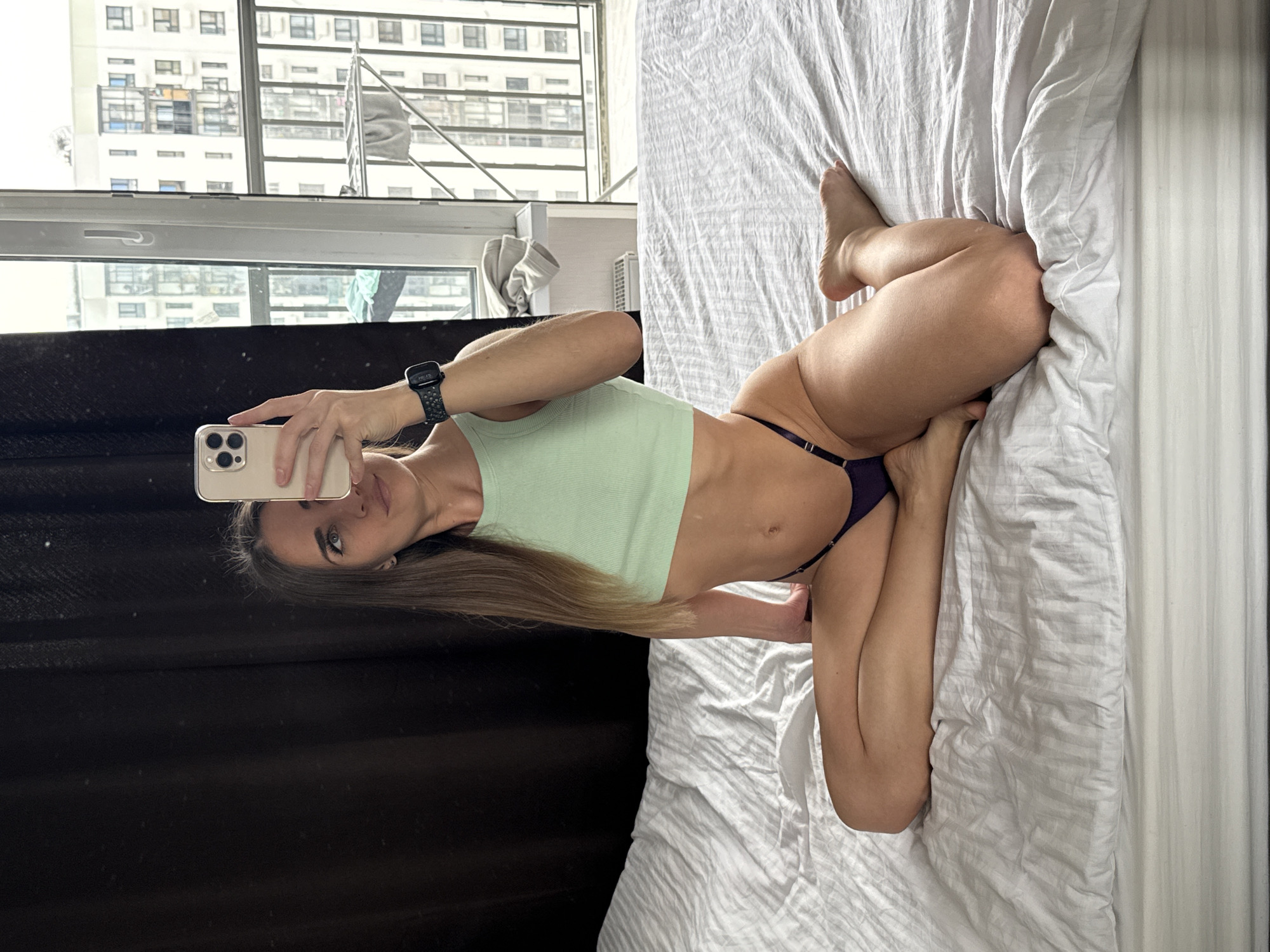 A woman is lying on a bed, taking a selfie with her smartphone. She's wearing a light green top and a dark purple thong. The bed has white sheets and is located in a room with a window showing buildings. Through the window, the cityscape of Lyubertsy, Russia can be seen. A dark curtain is visible to the left of the bed.