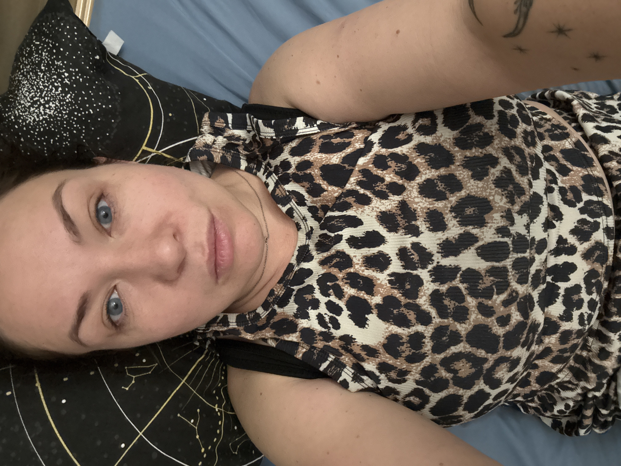 A woman with light skin and blue eyes is lying on a bed, propped up on a pillow with a black fabric cover featuring a celestial design in white and gold. She is wearing a leopard-print top with black straps. She has a small tattoo of a moon and stars on her right arm. The bed has a blue sheet. The location is unknown and there is no supporting information in the image.