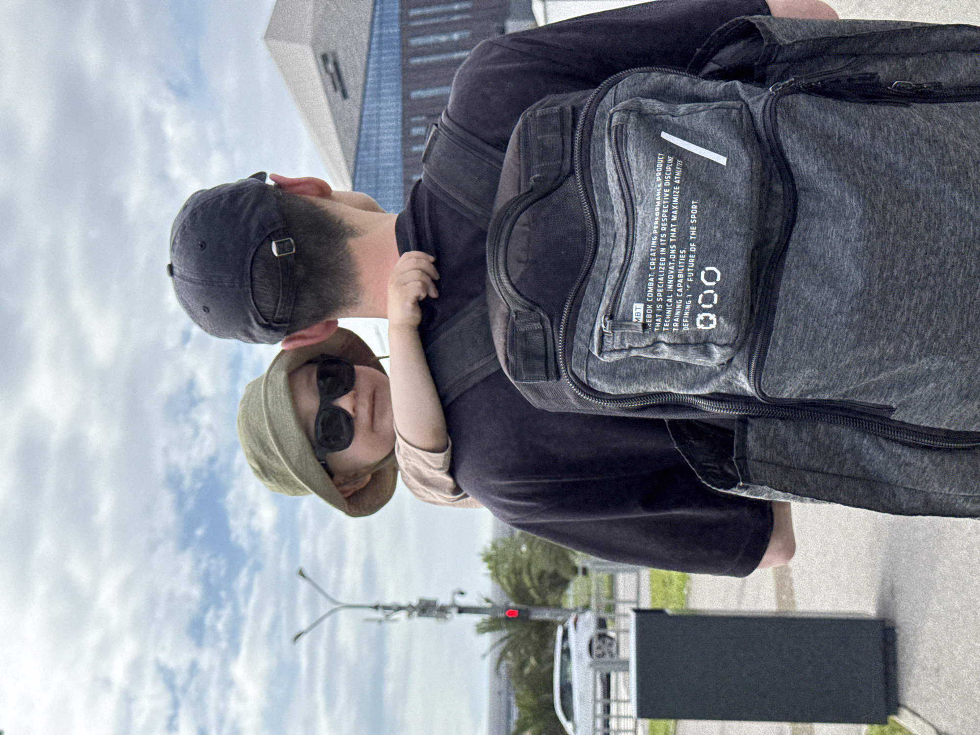 The image depicts a man carrying a young child on his back, with the scene set against an urban backdrop. The man is wearing a dark-colored top and a cap, and he has a gray backpack. The child is wearing a hat and sunglasses. The background includes a modern building with geometric shapes, a street with a white car, and a traffic light. The location is likely Sochi, Russia.