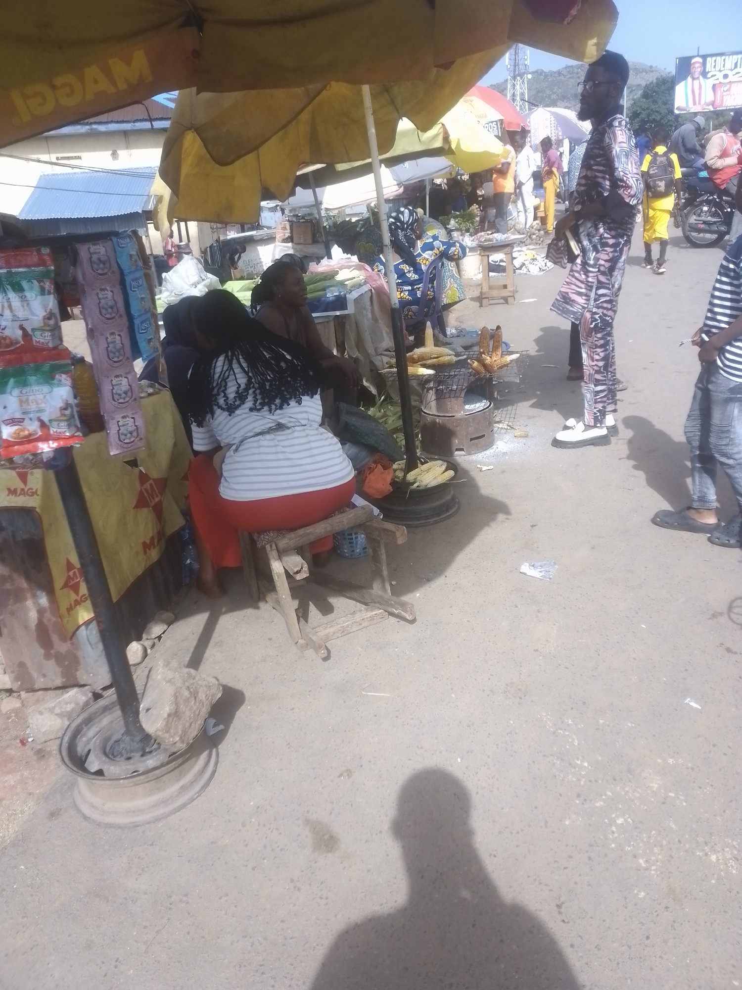 This media file captures a bustling outdoor market scene, likely in a warm climate given the bright sunlight and shadows. The primary subjects are several people engaged in various activities typical of a market. In the foreground, a woman is seated on a stool, her back to the camera, with her hair in braids. She appears to be part of the market stall, possibly selling or preparing food. Nearby, another woman is visible, seemingly interacting with the first.
The scene is set under a large, yellow umbrella that provides shade. In front of the seated woman, a charcoal grill is set up, with several ears of corn roasting on it, indicating that corn is being sold. There are also stacks of packaged goods, possibly food items or other merchandise, displayed on a shelf to the left. Some of these packages have visible text, including what appears to be "MAGGI" prominently featured on a yellow cloth banner. Further into the background, more people are milling about, and a motorcycle is parked. One individual, a man wearing a patterned outfit and glasses, stands out in the middle ground, facing away from the camera. Another man, in a striped shirt and jeans, is partially visible on the right.
The overall atmosphere suggests a busy daytime market. The bright sunlight casting distinct shadows indicates it's likely midday. There are no clear cues about the specific location, but the presence of a billboard with a politician's image ("REDE... 2023") suggests an election period and a populated area. The environment appears somewhat dusty and informal, characteristic of many street markets. The activity is centered around the food stall, with the preparation and sale of roasted corn being a key focus.
