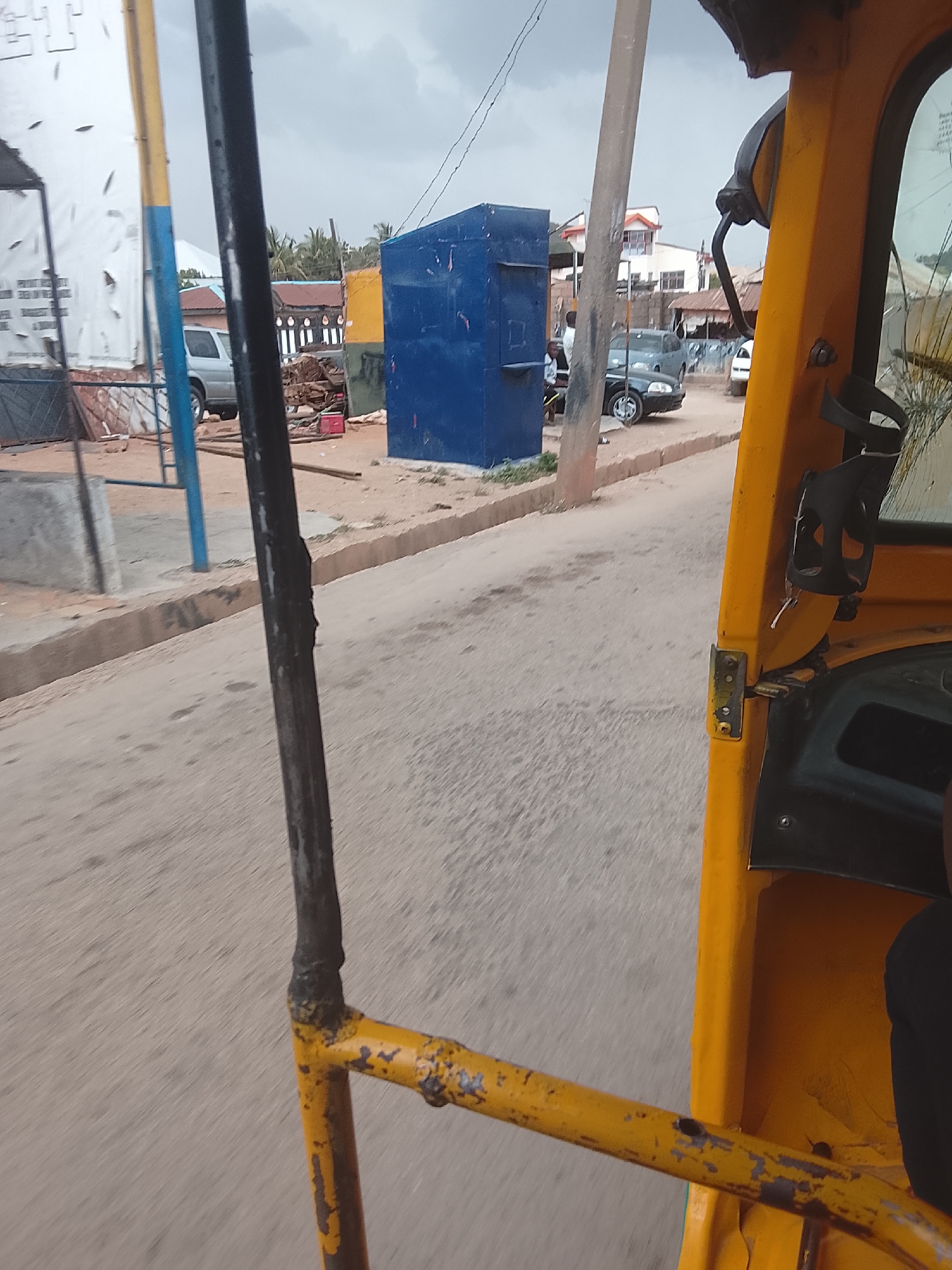 The image, taken from the interior of a yellow auto-rickshaw (keke) in Jos, Nigeria, shows a street scene. The camera appears to be positioned from the passenger seat. A blue, rectangular structure is visible on the side of the road. Several vehicles, including a silver SUV and a dark-colored car, are parked along the street. Other roadside features include utility poles and a partly visible white advertising board. The sky is overcast.