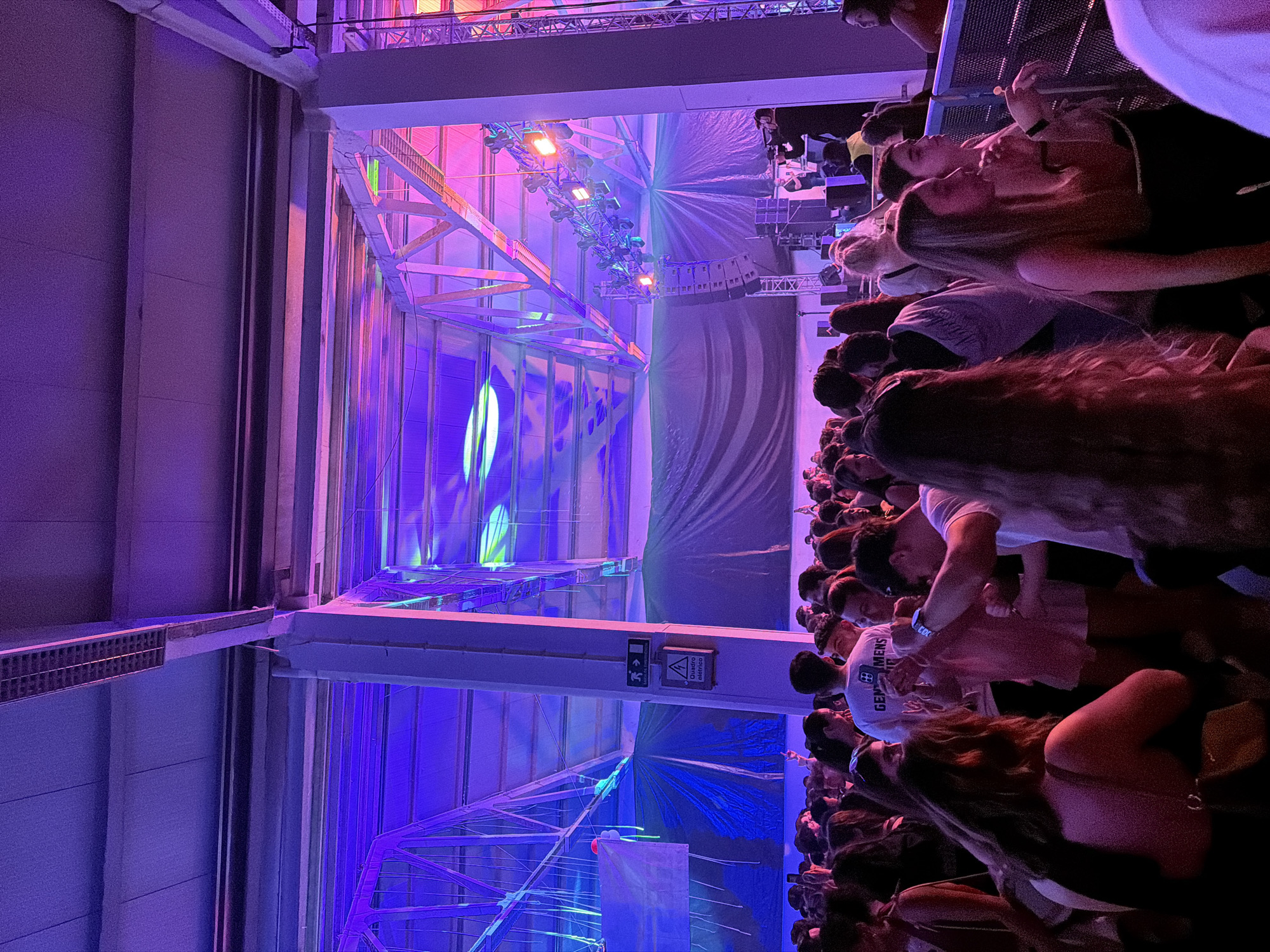 The image captures an energetic indoor event, likely a concert or party, held within a large hall in Bragança, Portugal. The venue is bathed in dramatic, artificial lighting, primarily vibrant purple and blue hues, with some orange and pink tones emanating from stage fixtures.
In the foreground and extending along the right side of the frame, a large crowd of attendees is visible, predominantly facing towards a distant stage. The faces and figures of the audience members are illuminated by the colorful lights, suggesting a lively and engaged atmosphere. One person in the crowd wears a white t-shirt with the word "GENTLEMEN'S" and a logo printed on it.
The central and left parts of the image reveal the structural elements of the building and the elaborate stage setup. Large, grey industrial beams and cross-sections of the hall's framework are prominent. Towards the rear, a complex array of metal trusses supports numerous professional lighting fixtures, which project powerful beams of colored light across the space. Behind these structures, large fabric backdrops or screens catch the light.
An overhead beam features a white sign with black text and symbols. It displays an exit symbol, indicating a path to the right and down. Below this, a triangular warning symbol accompanies the Portuguese text "quadro elétrico," which translates to "electrical panel." The event takes place at night or in the evening, given the reliance on artificial lighting.