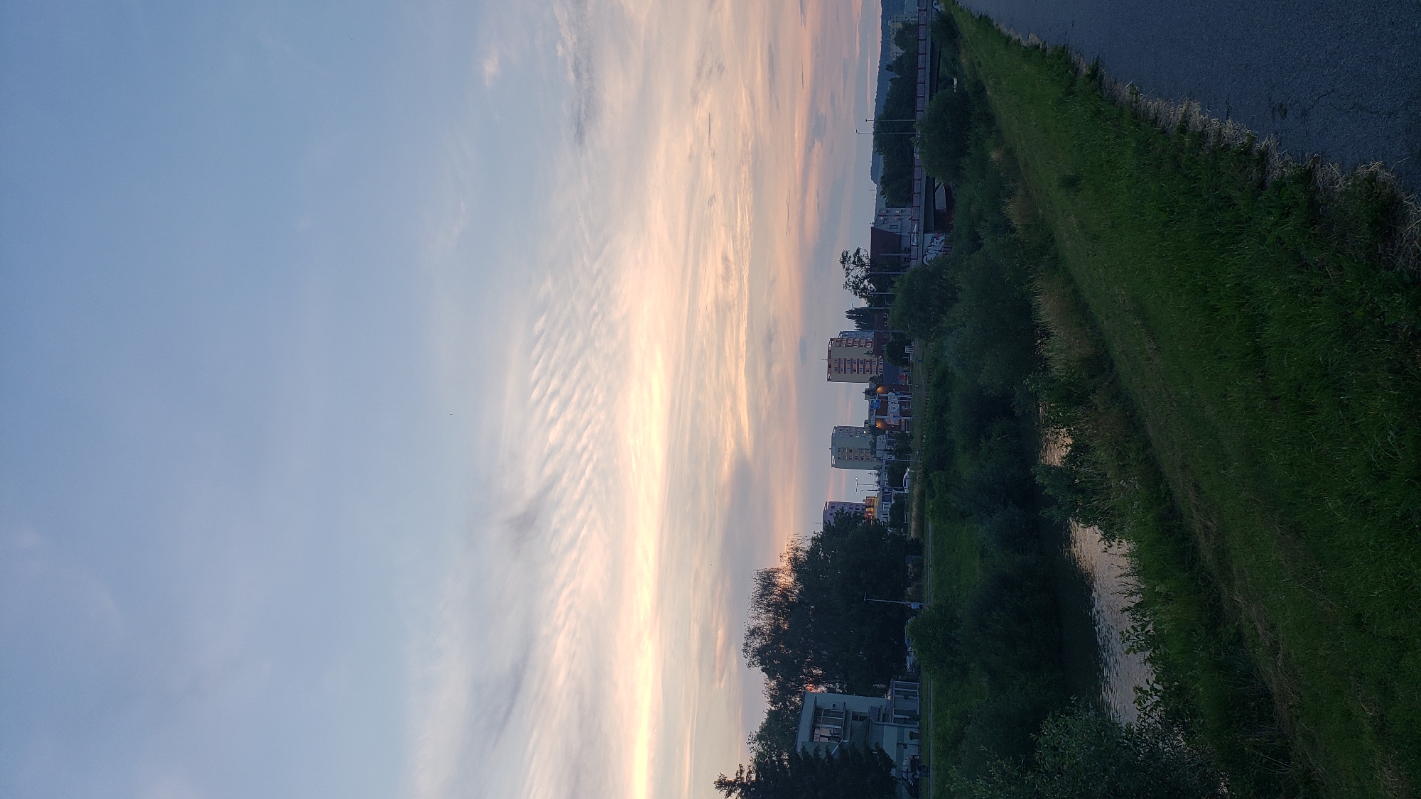 This image depicts a twilight scene in the city of Prešov, Slovakia, likely from the area near the Sekčov riverbank. The sky is a soft gradient of pale blue and pink, with delicate, wavy cloud formations catching the fading sunlight. In the middle ground, several multi-story buildings, typical of urban residential areas, are visible, some with colorful facades. Dense green foliage and trees line the river and the foreground, obscuring much of the ground level. A paved path, possibly a sidewalk or bike lane, runs along the right side of the frame, bordered by a grassy embankment. The river itself is a dark, reflective surface, mirroring the muted colors of the sky and vegetation. No people are clearly discernible in the image.
