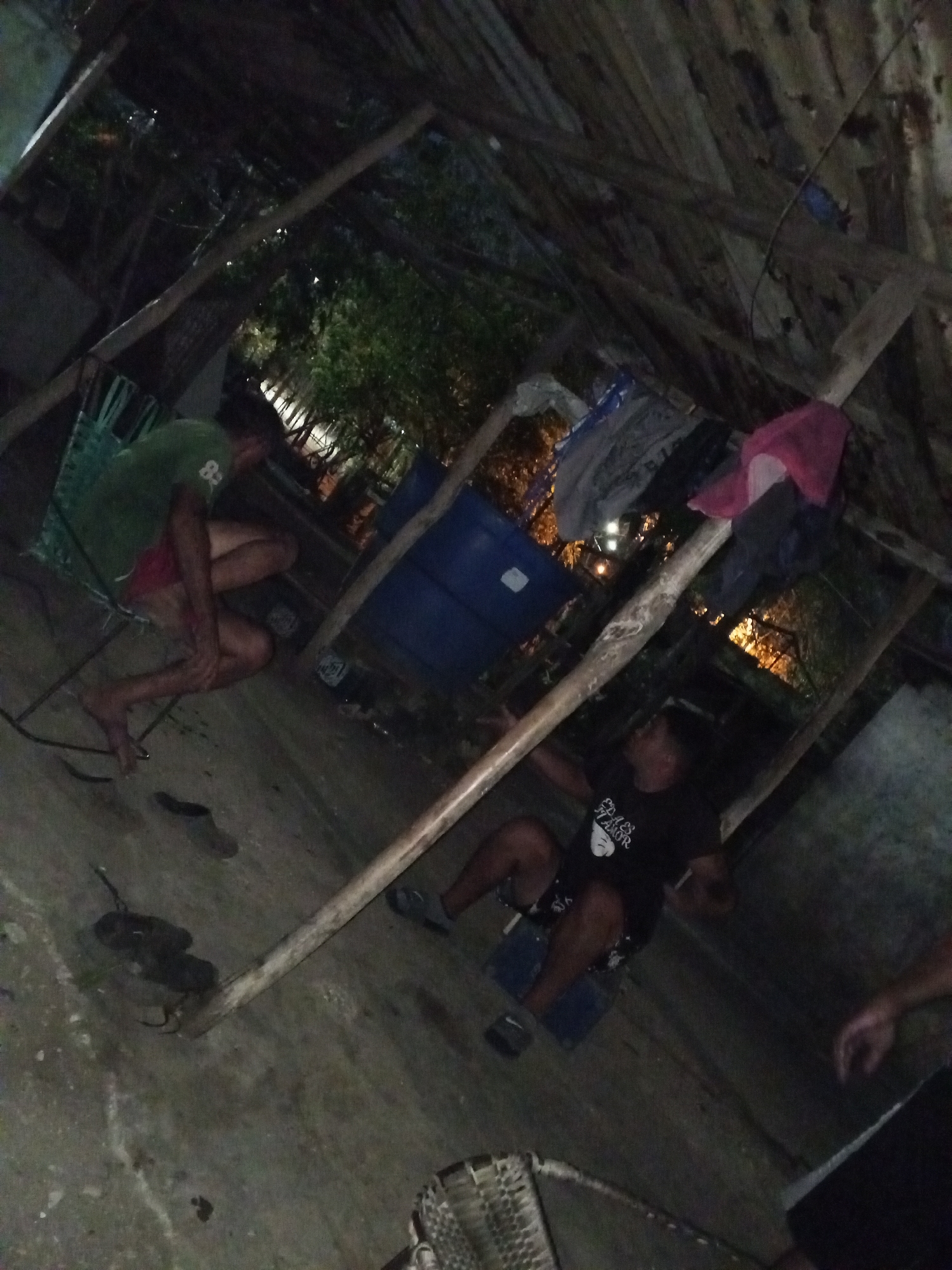 The image appears to be taken at night under some sort of ramshackle shelter or building frame in Altagracia, Venezuela. The structure is made of wooden beams and what looks like corrugated roofing. Clothing or fabric items are hanging from the structure. A blue drum sits in the background. The lighting is dim. There are at least two individuals, one wearing a green shirt and red shorts, the other a black t-shirt. Both individuals are sitting down. Several pairs of shoes are on the ground. Another person is standing in the foreground, cropped from the waist up. The ground appears to be concrete.