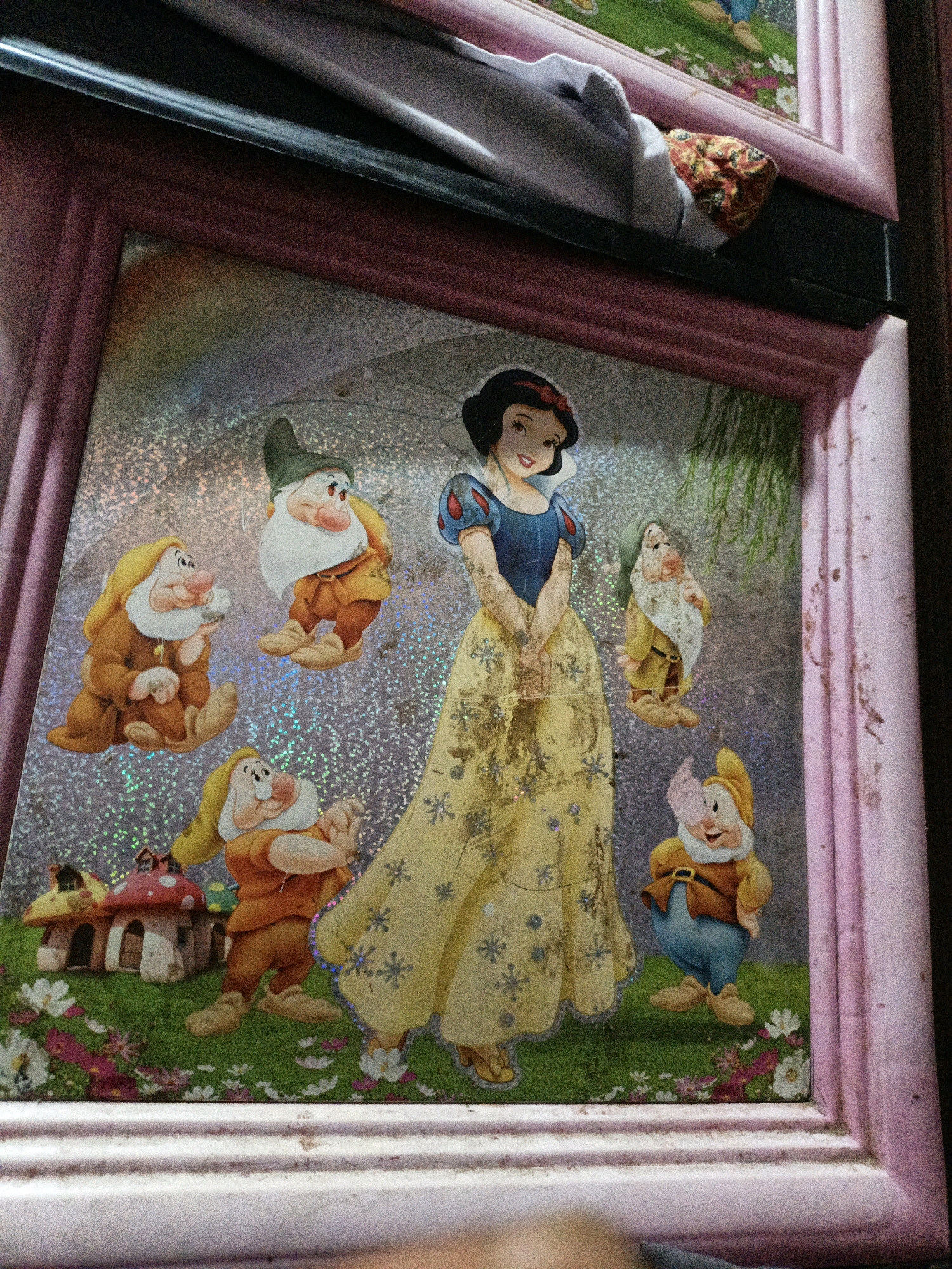 A framed image depicts a scene from Snow White and the Seven Dwarfs. Snow White stands in the center, wearing her iconic blue and yellow dress. Six of the seven dwarfs are arranged around her: Doc, Grumpy, Happy, Sleepy, Bashful, and Sneezy. A mushroom-shaped house is visible on the left side of the image. The background is a sparkly, silver material with hints of green foliage. The frame surrounding the image is pink and appears to be made of plastic. The image is leaning against a dark surface, and part of a patterned fabric is visible at the top. The scene is indoors, possibly in a home in West Jakarta, Indonesia.