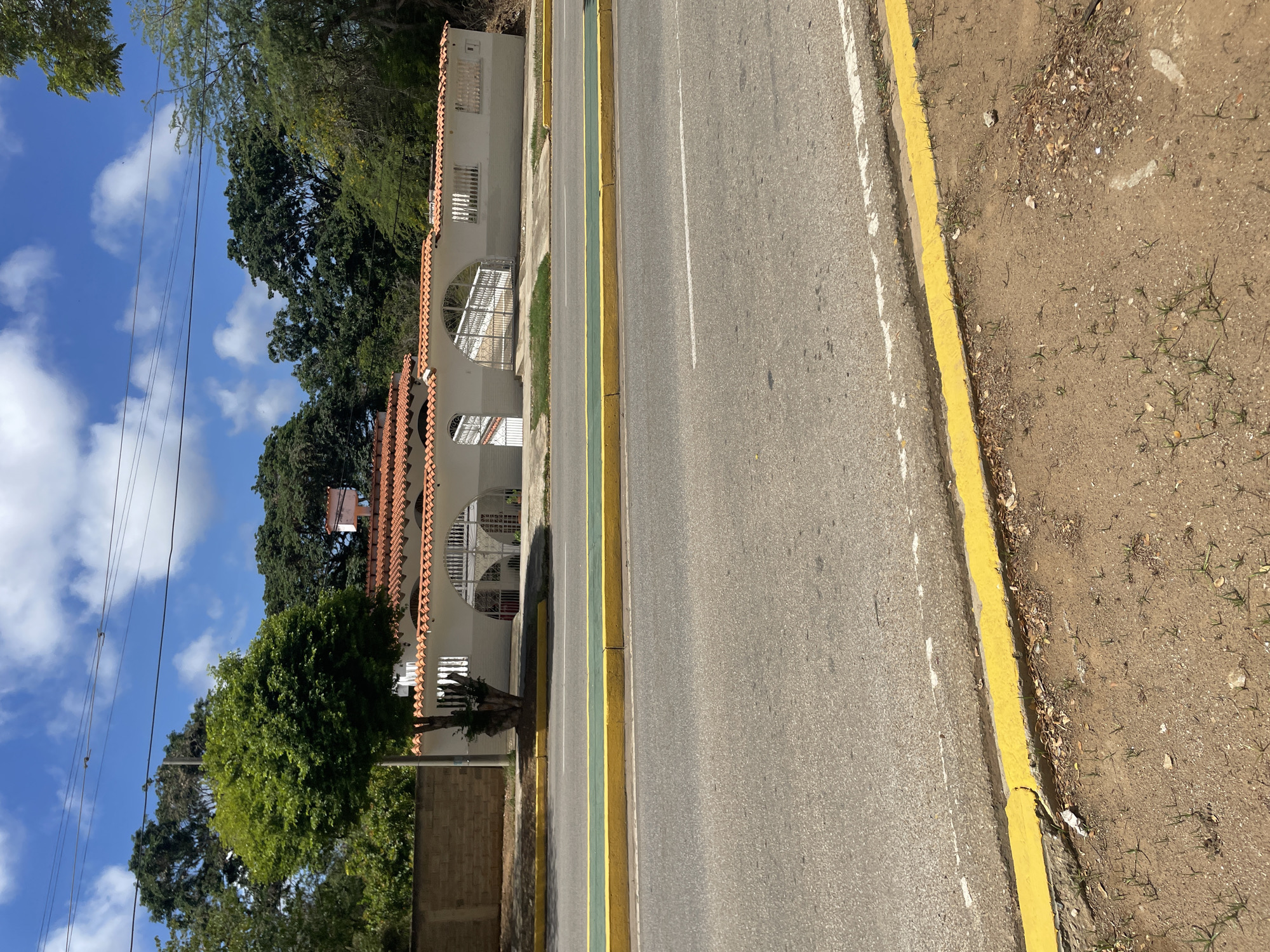 The image shows a street scene in El Valle del Espíritu Santo, Venezuela. A white building with a terracotta tile roof and decorative arched entrances is visible behind a low wall. The building appears to be a residence or a commercial establishment. The street in front of the building is paved and has yellow and green painted curbs. Trees and foliage surround the building and extend into the background. The sky is blue with scattered white clouds. There are no people visible in the image. The scene appears to be tranquil and bathed in sunlight.