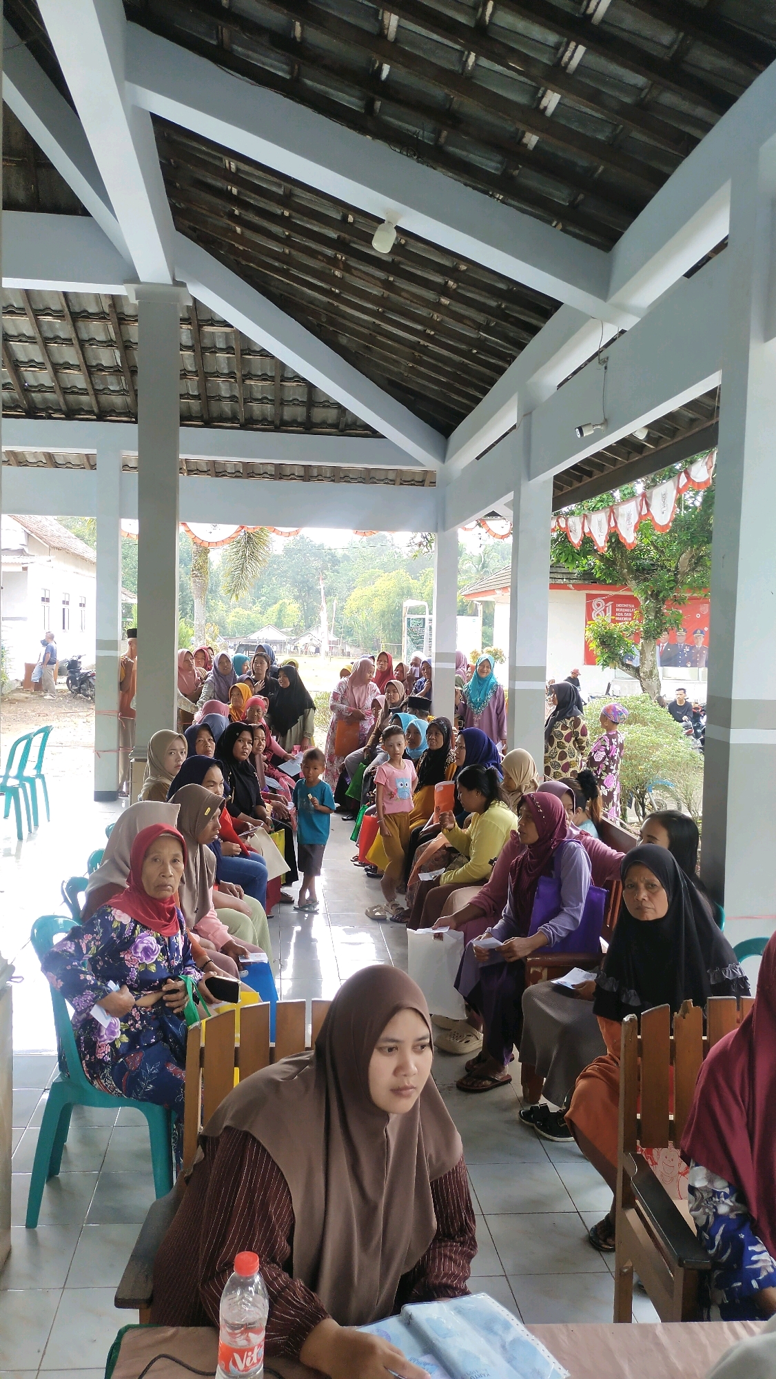 An outdoor, covered waiting area is filled with approximately 30 Indonesian women and a few children. They are seated on plastic and wooden chairs and appear to be waiting for an event or service. Most of the women are wearing hijabs and traditional Indonesian clothing. In the foreground, a woman in a brown hijab is seated at a table with a bottle of water and some documents. The background shows trees and buildings, suggesting a rural or semi-rural setting in Klungkung, Indonesia. The architecture of the waiting area, with its exposed wooden roof beams and white pillars, indicates a functional, open-air structure. The overall atmosphere seems calm and orderly.