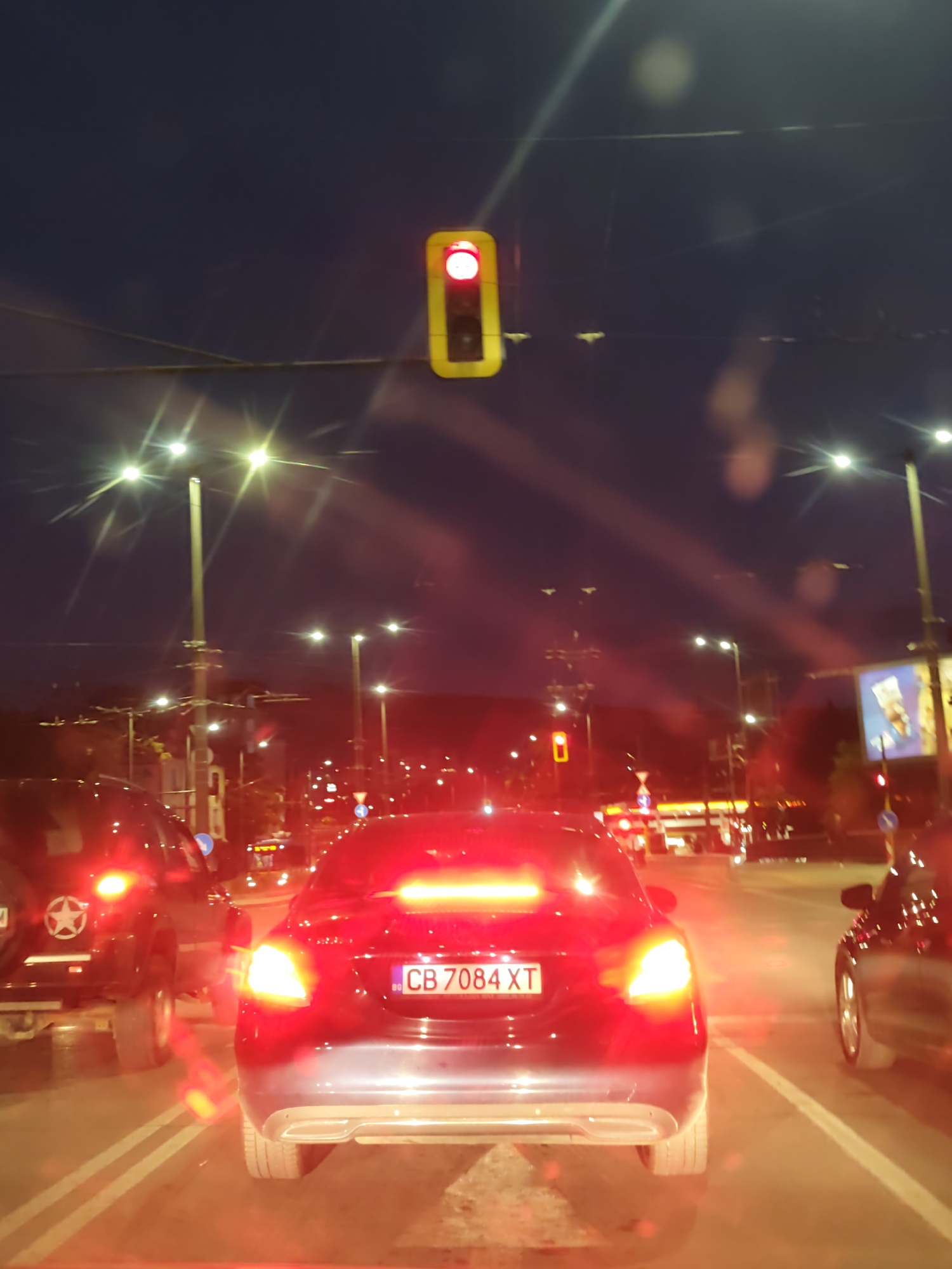 The image depicts a nighttime scene on a street in Sofia, Bulgaria, likely during heavy traffic. Several cars are visible, illuminated by their brake lights, including a dark-colored SUV with a star insignia on its rear. The car in the foreground has a Bulgarian license plate "CB 7084 XT." Streetlights and a traffic light displaying red are present, indicating the presence of an intersection. A fuel station is visible in the distance. The street is marked with white lines, delineating lanes. The sky is a dark gradient blue, suggesting twilight.