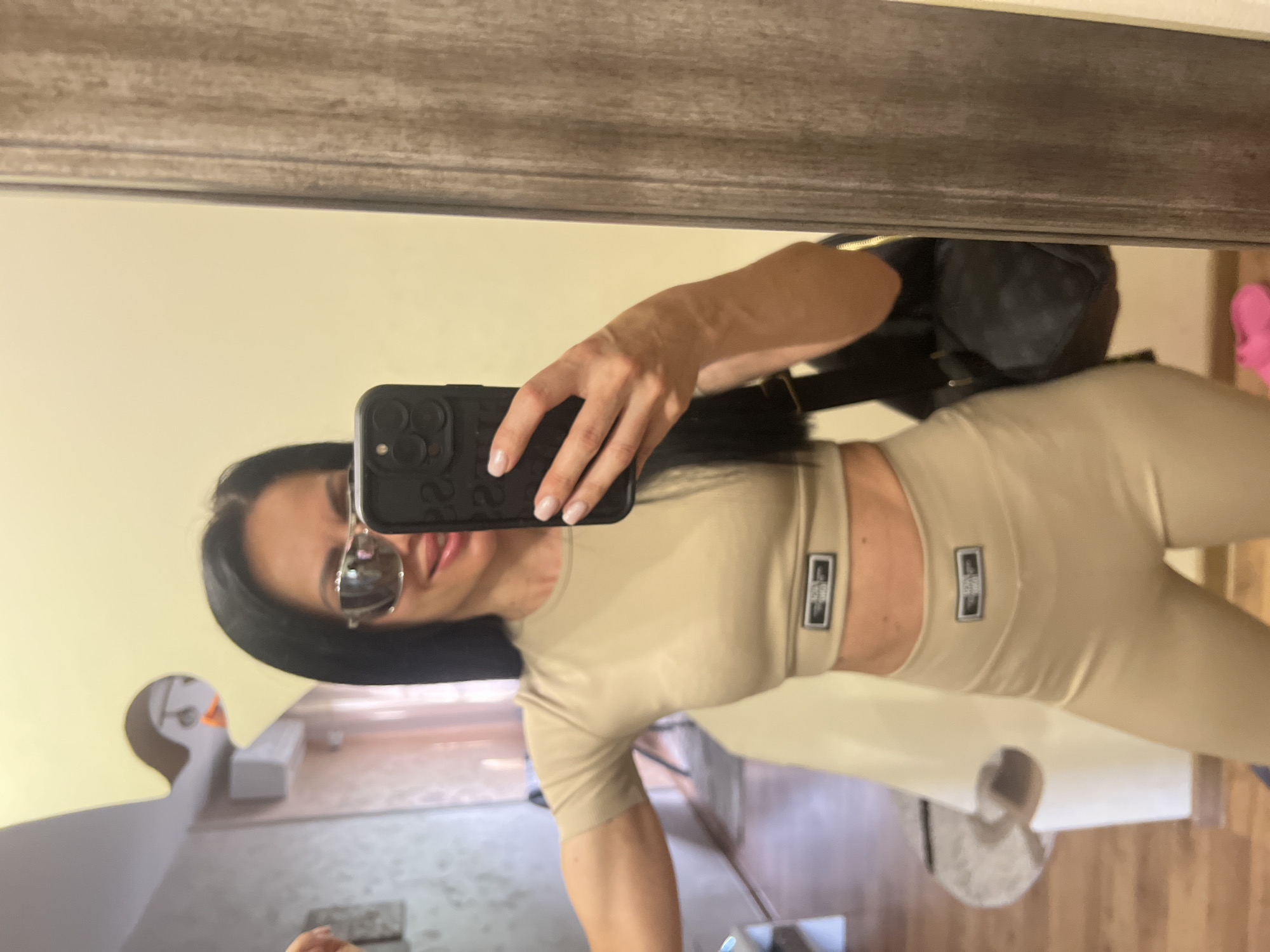 A woman is taking a mirror selfie in what appears to be a residential interior. She has long, dark hair, is wearing large sunglasses, and a beige crop top and matching leggings with logos on them. She is holding a smartphone in her right hand, which is capturing the reflection. A dark handbag is slung over her left shoulder. The background shows a wall with a mirror, a doorway with a wooden frame, and part of a room with furniture. The mirror has a unique puzzle-piece shaped cutout in the upper left corner. There is no information that can be used to determine the location in Stara Zagora, Bulgaria.