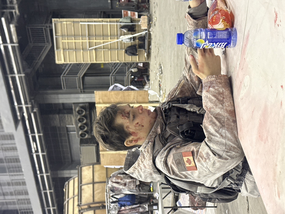 A young individual with short light brown hair exhibits simulated lacerations and extensive bloodstains on the face, specifically across the forehead, left cheek, and around the nose/mouth area. The individual is seated at a light-colored table, which is also marked with numerous red liquid stains. They wear a camouflage-patterned hooded jacket and a tactical vest, both heavily splattered with red liquid. A patch depicting the Canadian flag and an additional emblem is visible on the left sleeve. A dark-colored backpack with grey straps is worn. The individual's gaze is directed downward towards an item held in their hands, which is largely obscured.
On the table, a blue plastic bottle with a blue cap and a label featuring Chinese characters and a yellow/blue graphic is present, alongside a translucent red-orange bag, possibly containing snacks.
The background depicts a large, high-ceilinged indoor space, characteristic of a film studio or industrial facility. Architectural elements include exposed metal structures, multiple ventilation units, and multi-level metal staircases with railings. To the immediate left, a clothing rack holds numerous garments, many appearing blood-stained, next to a blue backpack. Various wooden set pieces, including tall flats and a larger structure, are visible further back. Debris is scattered across the concrete floor. The setting is a large indoor facility in China, evidenced by the Chinese characters on the beverage bottle; however, the precise location within Huangdao District is not visually identifiable.