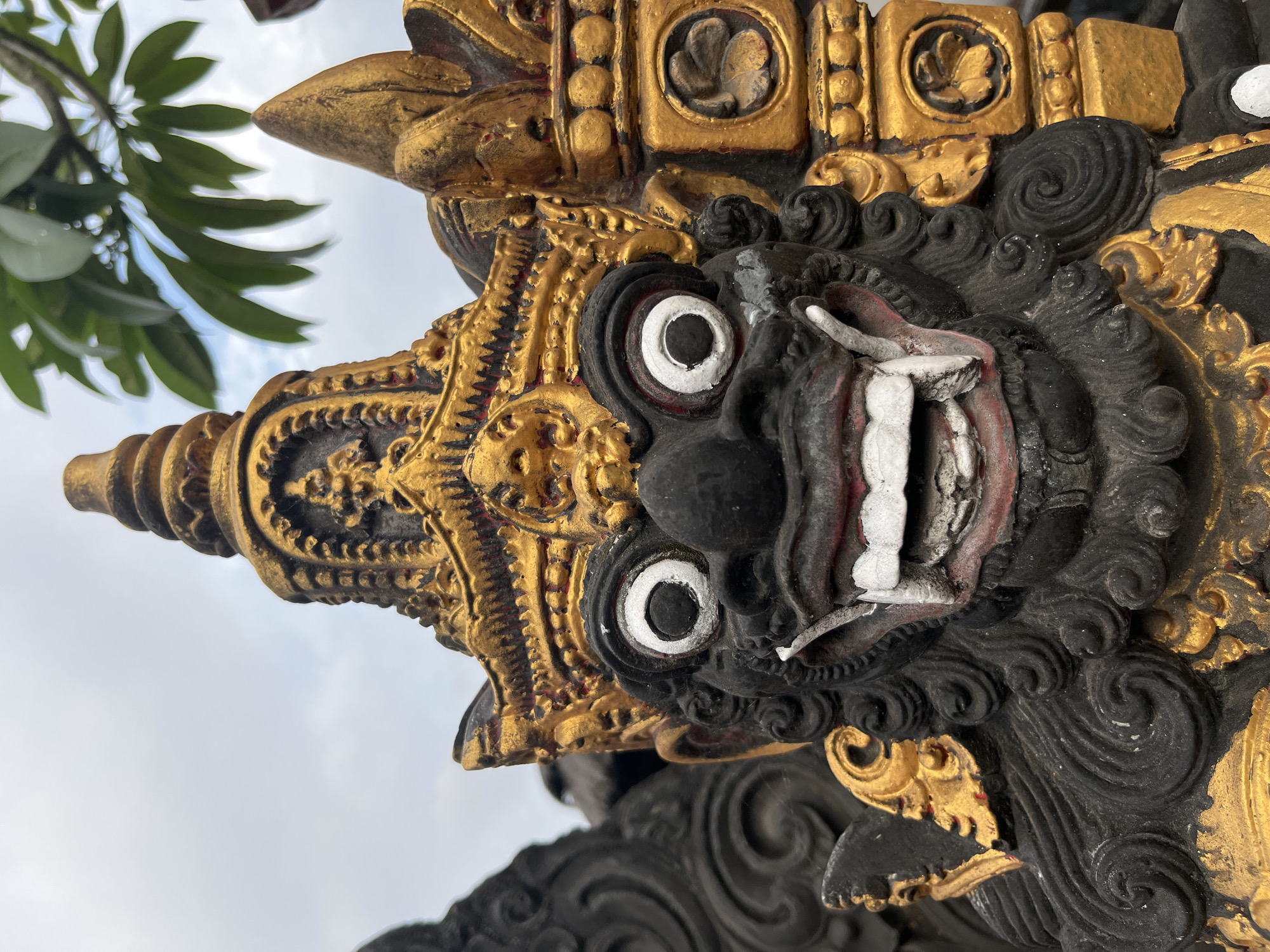 An extreme close-up shot focuses on the face of a black and gold Balinese statue, likely a demon or guardian figure, in Kuta Selatan, Indonesia. The statue has wide, staring white eyes with black pupils, a gaping mouth revealing sharp white teeth and red gums, and a black, textured face. It wears an elaborate gold headdress adorned with red and black details. To the right of the statue's face is a gold-colored decorative pillar with carved circular motifs. In the background, a cloudy sky and lush green leaves are visible.
