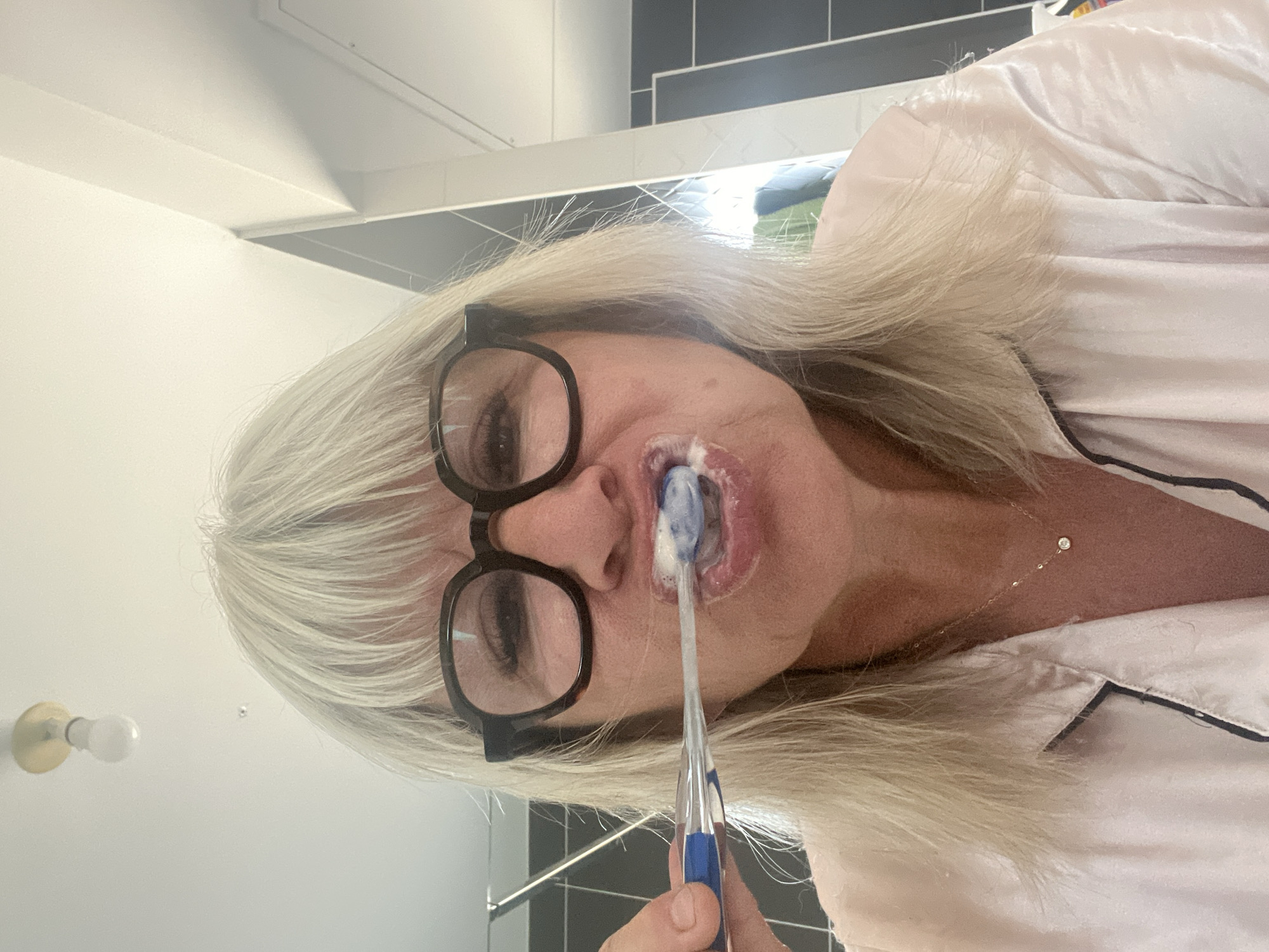 A medium shot captures a woman with blonde hair and bangs, wearing black-rimmed glasses and a light pink satin pajama top, brushing her teeth in what appears to be a bathroom. She is holding a blue and clear toothbrush with toothpaste in her mouth. The background features a white ceiling with a light fixture, a wall with grey and white tiles, and a glimpse of a shower curtain or door. The image's lighting is bright and direct, suggesting an indoor setting. The woman is looking directly at the camera, mid-action. There is no indication of the image being located in Chambœuf, France.