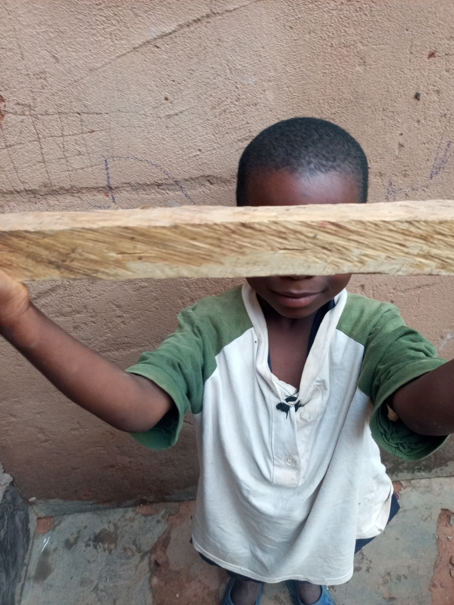The image depicts a young Black child, likely a boy, standing outdoors against a textured, light brown wall. The child is wearing a short-sleeved shirt with white and green panels. A horizontal piece of rough-hewn wood is held in front of the child's face, obscuring their eyes and nose. The child's arms are bent, holding the wood. The background wall shows faint markings, possibly graffiti or natural wear. The ground beneath the child appears to be concrete or a similar rough material with some debris. The overall setting suggests an informal, possibly rural or urban fringe, environment within Tempure, Nigeria.