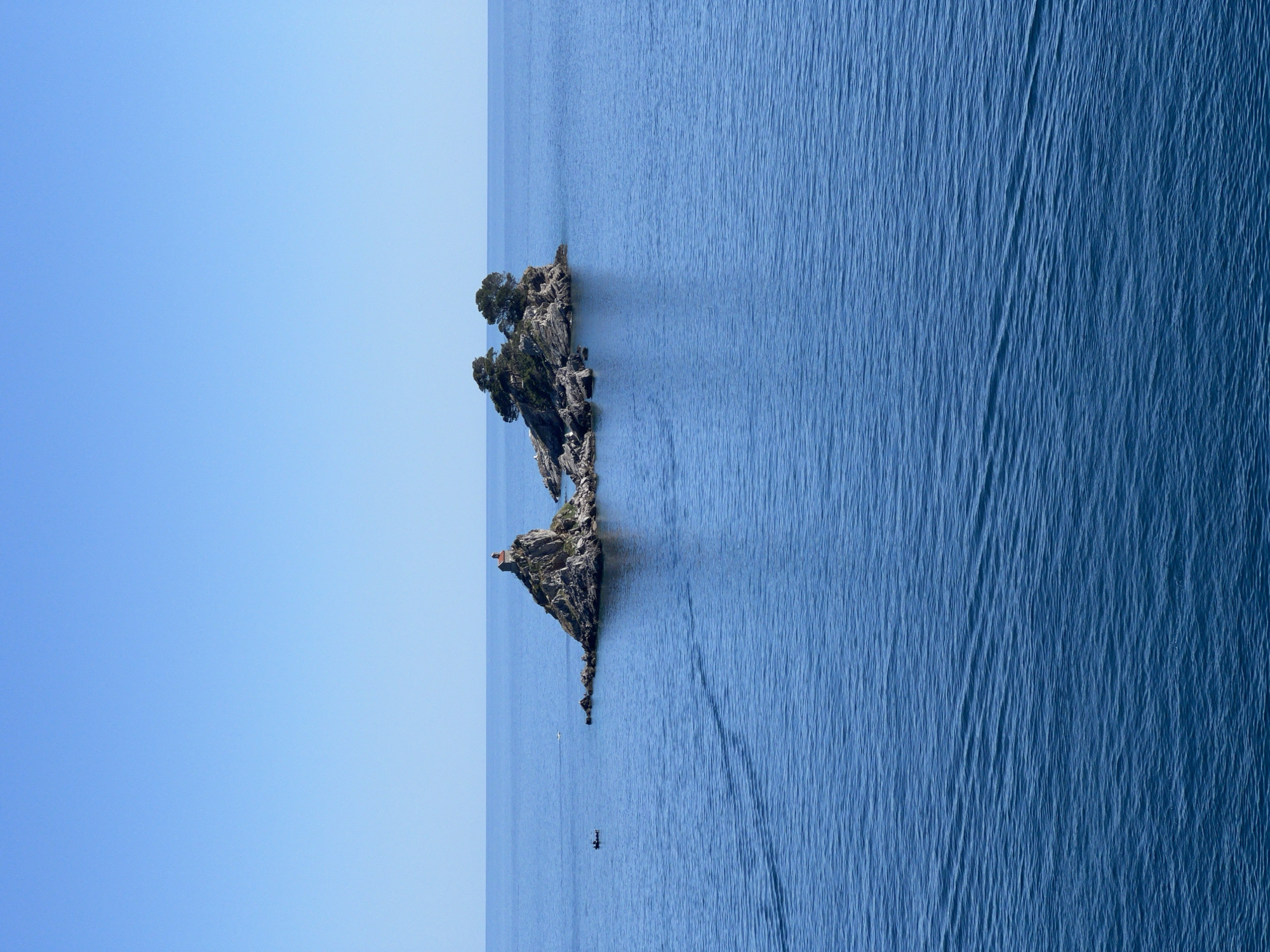 This vertically oriented image, best viewed rotated 90 degrees clockwise to a horizontal landscape, depicts a tranquil coastal scene under a clear, bright sky.
A prominent rocky island, covered with green trees and sparse vegetation, rises from the deep blue sea. The island occupies the left portion of the frame, tapering into a narrow rocky spit that extends further into the water. A small, light-colored structure, possibly a building or shrine, is perched on one of the higher points of the island.
The expansive sea fills the right side and bottom of the frame, characterized by gentle ripples and subtle wave patterns on its surface, indicating calm weather. The water exhibits a gradient of blue, becoming richer and deeper further from the horizon.
In the water near the island's tip, a small white boat or kayak is visible, along with a darker, indistinct figure, possibly a person, nearby, suggesting human presence or activity on the water. A dark, elongated shadow stretches across the water from the island.
The sky, a uniform gradient of clear light blue, dominates the upper portion of the image, entirely free of clouds. The bright daylight illuminates the scene, casting soft shadows. The overall impression is one of natural beauty and serenity, characteristic of a coastal landscape like Petrovac na Moru, Montenegro. There is no visible text in the image.