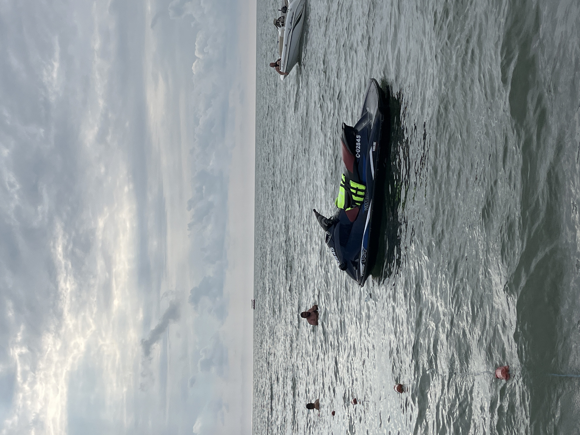 In the city of Ureki, Georgia, on a body of water, a jet ski is seen floating on the surface. The jet ski is dark blue with some red and yellow accents. In the background, a white boat with several people on board is visible. Closer to the shore, there are three individuals in the water, appearing to be swimming or wading. Red buoys are scattered on the water, connected by a rope. The sky is overcast with scattered clouds, and the water is calm with small waves. The overall scene suggests a recreational day at the beach.