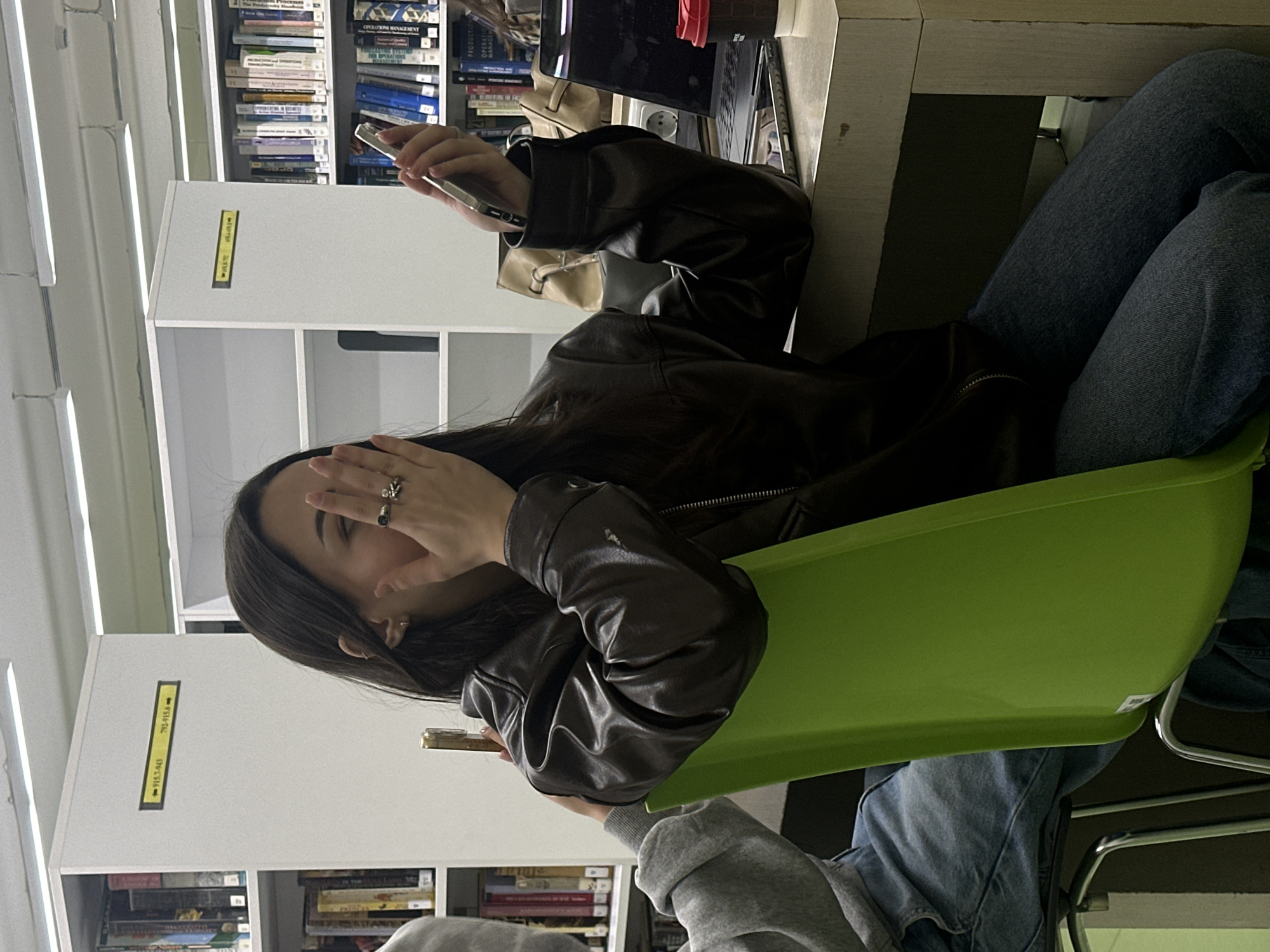 A woman is seated at a table in what appears to be a library, likely in Yerevan, Armenia, given the potential location request. She is wearing jeans and a brown leather jacket, sitting in a bright green chair. She holds a cell phone in one hand and her other hand is raised towards her face, covering part of it. Her dark hair is loose. The background shows bookshelves filled with books and a laptop is on the table.