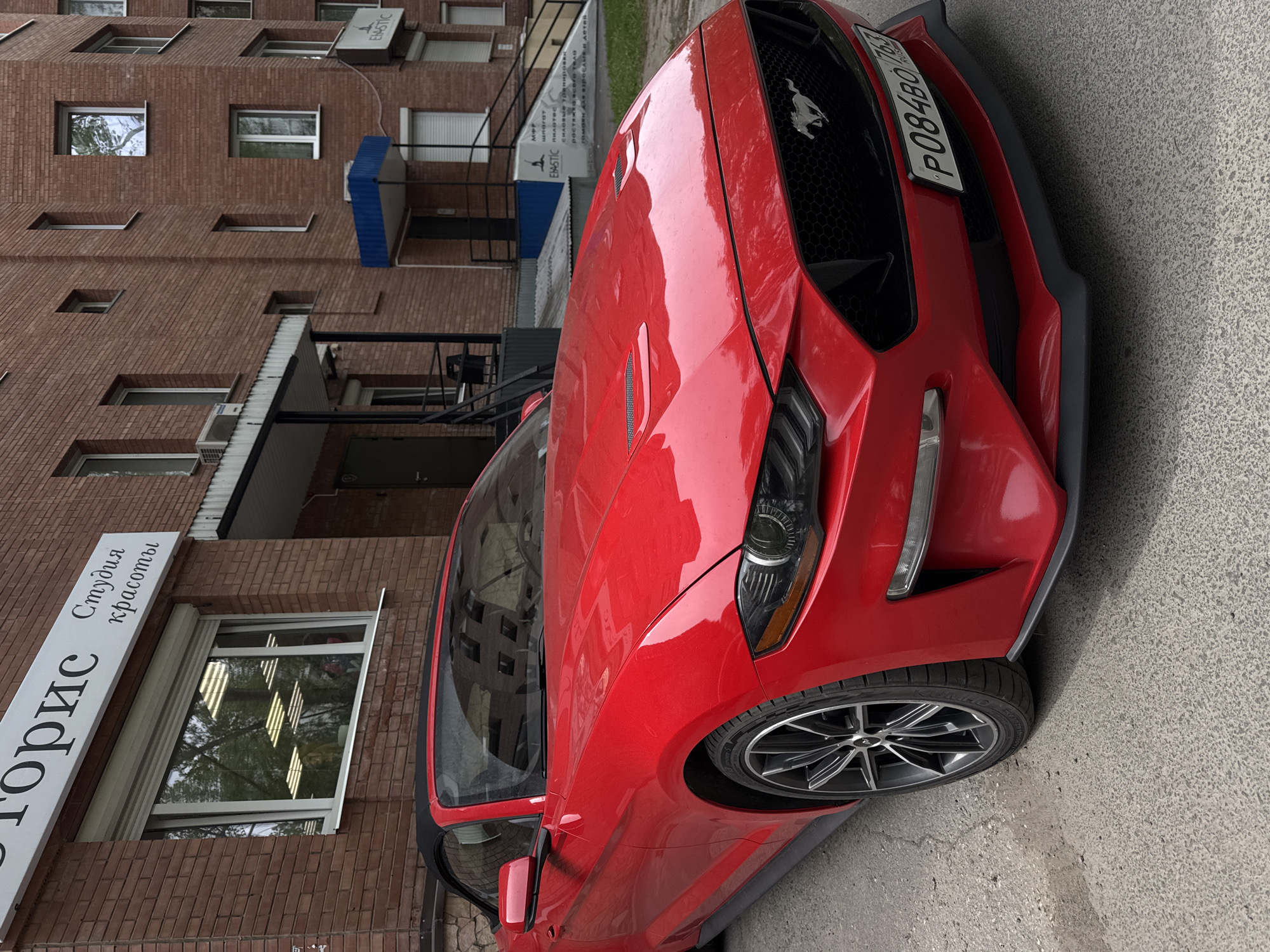 A red Ford Mustang is parked on a gray asphalt road in front of a brick building. The building has multiple windows and signs with Russian text. The car's license plate is visible. The day appears to be overcast.