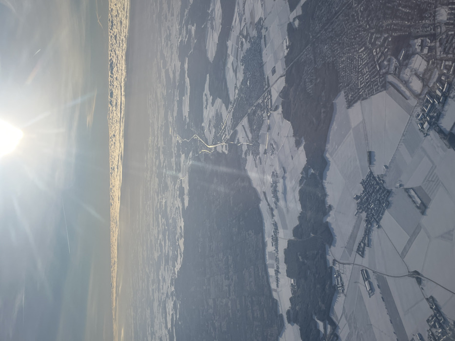 An aerial photograph captures a snow-covered winter landscape, likely taken from an aircraft. The sun, prominent in the upper right, casts intense flares and streaking rays across the scene. The sky above is clear blue, while a distinct, elongated band of stratified clouds spans the distant horizon, approximately one-third of the way up from the bottom edge. A faint contrail is visible in the upper left quadrant.
The terrain below is extensively covered in snow, appearing white and textured, with darker areas indicating exposed land or dense forestation. A prominent river, the Isar River, flows from the top-middle of the frame towards the bottom-right, visible as a darker, winding line against the snow. Parallel to the Isar River on its western bank, a major highway (likely Bundesstraße 11 or Autobahn 92) traverses the landscape, appearing as a bright, linear feature reflecting sunlight.
The city of Freising, Germany, is identifiable in the bottom-right quadrant of the image as a sprawling urban area with a high density of buildings and a discernible street network. Buildings and rooftops within Freising are covered in snow. Smaller villages and hamlets, characterized by clusters of structures amidst snow-covered agricultural fields, are scattered throughout the mid-ground and foreground. Patches of dark green/brown forests punctuate the landscape, contrasting with the white snow. No individuals are discernible within the frame due to the high altitude.