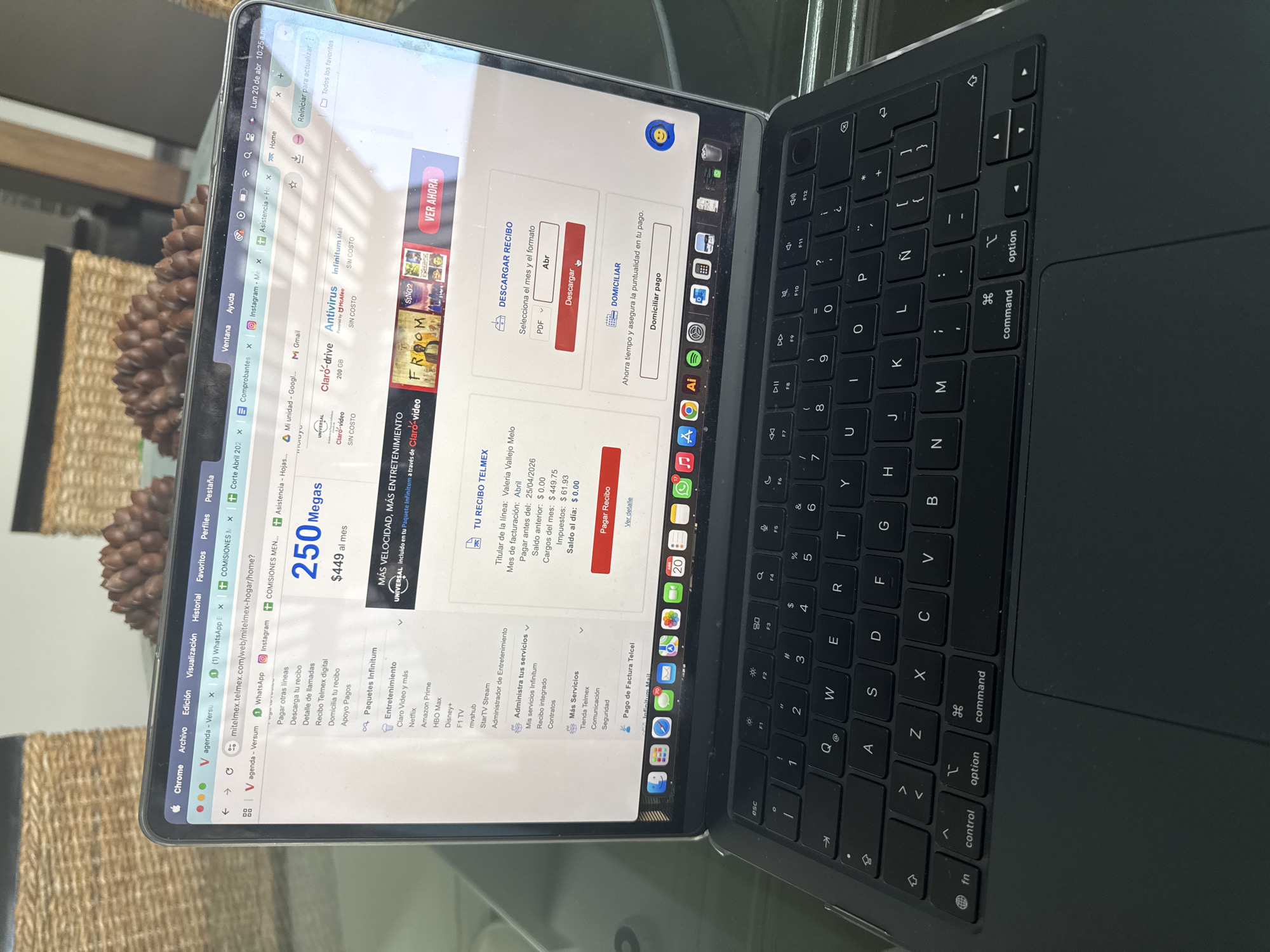 An open MacBook laptop, likely a Pro or Air model, rests on a glass surface, displaying a Telmex telecommunications service webpage. The scene suggests an indoor setting, possibly on a table, with a woven basket and a pinecone-like decorative object visible in the background.
The laptop screen, captured on Monday, April 20th, at 10:25 AM, shows a personal account page on mitelmex.telmex.com. The primary focus is a bill for "Valeria Vallejo Melo" for April, showing a previous balance of $0.00, current charges of $449.75, taxes of $61.93, and a present balance of $0.00, with a payment due date of "25/04/2026". The user's mouse cursor hovers over a red "Descargar" (Download) button, indicating an intention to download the bill in PDF format. Options to "Pagar Recibo" (Pay Bill) and "Domiciliar" (Direct Debit) are also prominent.
Additional details on the webpage highlight the service plan, advertising "250 Megas" for "$449 al mes" (per month). Bundled services such as "Claro-video", "Claro-drive (200 GB)", "Antivirus (Powered by McAfee)", and "Infinitum Mail" are listed as "SIN COSTO" (No Cost). A section for entertainment options includes Netflix, Amazon Prime, HBO Max, Disney+, F1 TV, mvshub, and StarTV Stream.
The laptop's black QWERTY keyboard with Spanish characters (like 'Ñ') and trackpad are visible. The operating system's dock shows various application icons, including Chrome, Safari, Mail, Messages, Calendar, Photos, FaceTime, Maps, Reminders, Music, App Store, Settings, Adobe Illustrator, Adobe Acrobat, Spotify, Downloads, and Trash. The overall activity depicts a person managing their Telmex bill online in San Bernardino Tlaxcalancingo, Mexico.