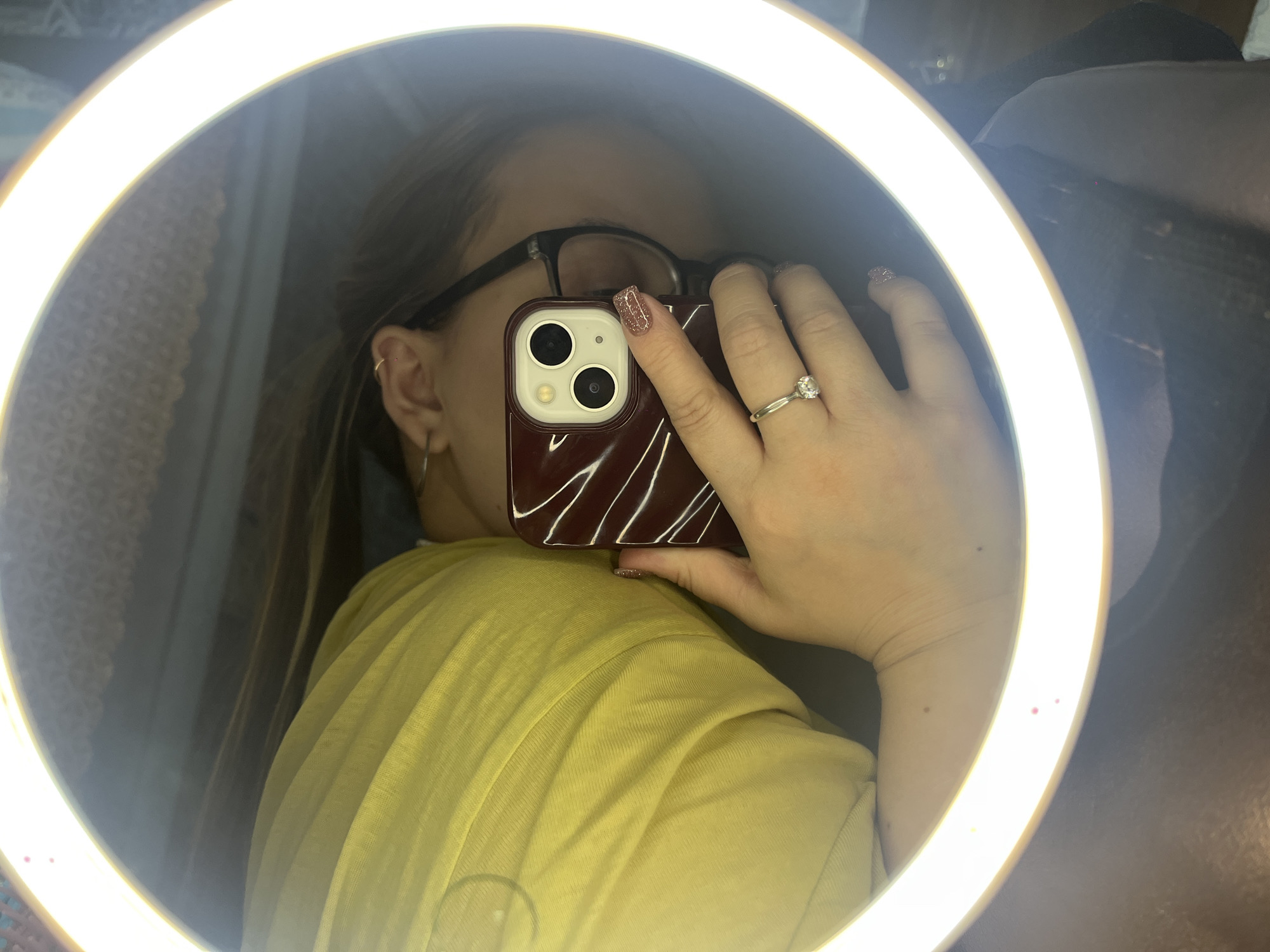 A person is taking a selfie in front of a ring light. The individual is wearing glasses and a yellow shirt. They are holding a cell phone with a red case and two lenses on the back, and a ring is visible on their finger. The surrounding environment includes curtains and what appears to be clothing. The location, Novopokrovskaya, Russia, is not discernible from the image.