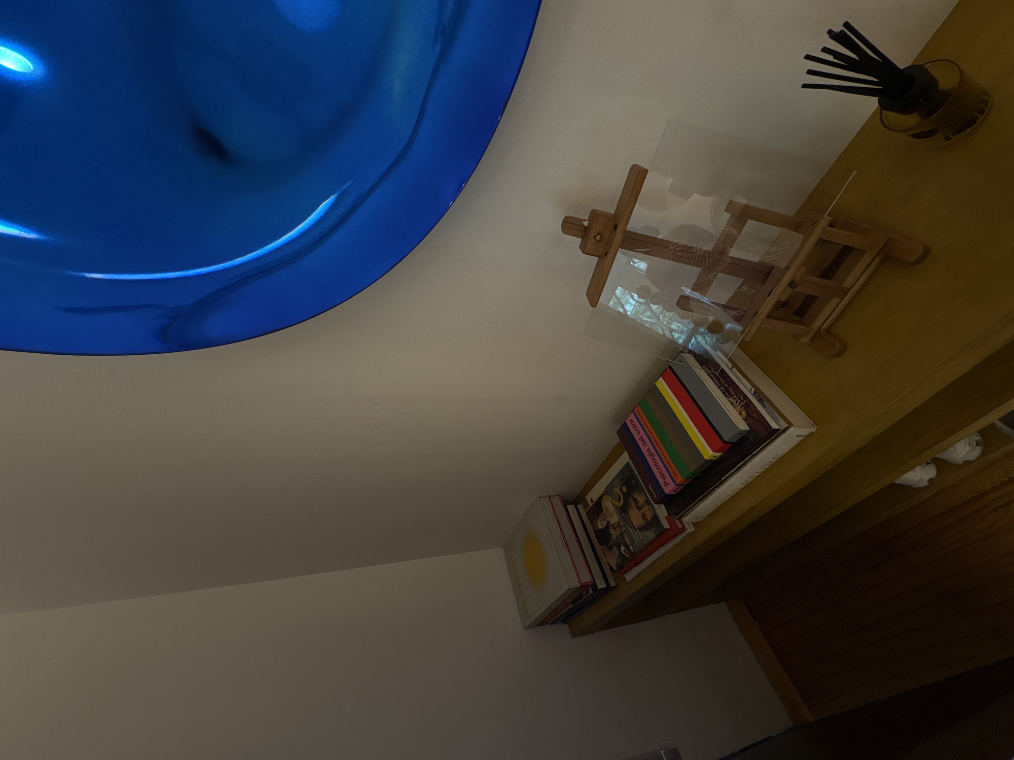 This image depicts an indoor scene in a room located in Mexico City, Mexico. The perspective is slightly elevated, looking down into a corner. A large, circular, convex art piece in a highly reflective, vibrant blue hue occupies the upper right portion of the frame, mounted against a light-colored wall. Its glossy surface shows distorted reflections of the surroundings.
Below and to the left of the blue art piece, a horizontal wooden shelf, light brown or yellow-toned, extends along the wall. On the far left of the shelf, a stack of books is visible; the top book has a white cover featuring a prominent yellow circular design. To its right, another stack of books includes a volume with a partial image resembling the Mona Lisa on its cover and text that appears to include "Esthetics." Beneath this, a book titled "Psicología del color" (Psychology of Color) with a multi-colored spine (featuring red, yellow, green, blue, brown, gray, and black segments) is clearly identifiable.
Further to the right on the shelf, a small, light-colored wooden artist's easel stands with a clear, rectangular acrylic or glass sheet leaning against its support. The sheet reflects patterns, possibly from an external window or light source. To the far right on the shelf, a dark brown or amber-colored cylindrical diffuser bottle contains several black reed sticks.
The walls are painted a neutral, light off-white color. The floor in the lower left corner consists of dark wooden planks. The lighting in the scene appears to be dim to moderate, casting soft shadows. There are no people present in the image.