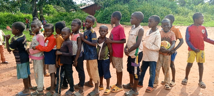 The image displays a group of approximately 15 children, primarily males, ranging in age from approximately 5 to 12 years old, standing on a reddish-brown dirt path in Ngalula, Democratic Republic of the Congo. The children are arranged in a loose line formation, generally facing towards the viewer's left. Their attire consists of assorted casual clothing, including t-shirts, long-sleeved shirts, shorts, and pants, with several wearing sandals or flip-flops. One boy, positioned towards the center-right of the frame, holds a yellow and black patterned soccer ball in his hands and exhibits a broad smile. The other children display mostly neutral or curious expressions. The immediate ground surface is unpaved, composed of dirt and appearing somewhat uneven. The background is characterized by dense green foliage, including trees and bushes, with a light-colored building topped by a dark roof partially visible through the vegetation on the left side. The scene is illuminated by bright, natural daylight.