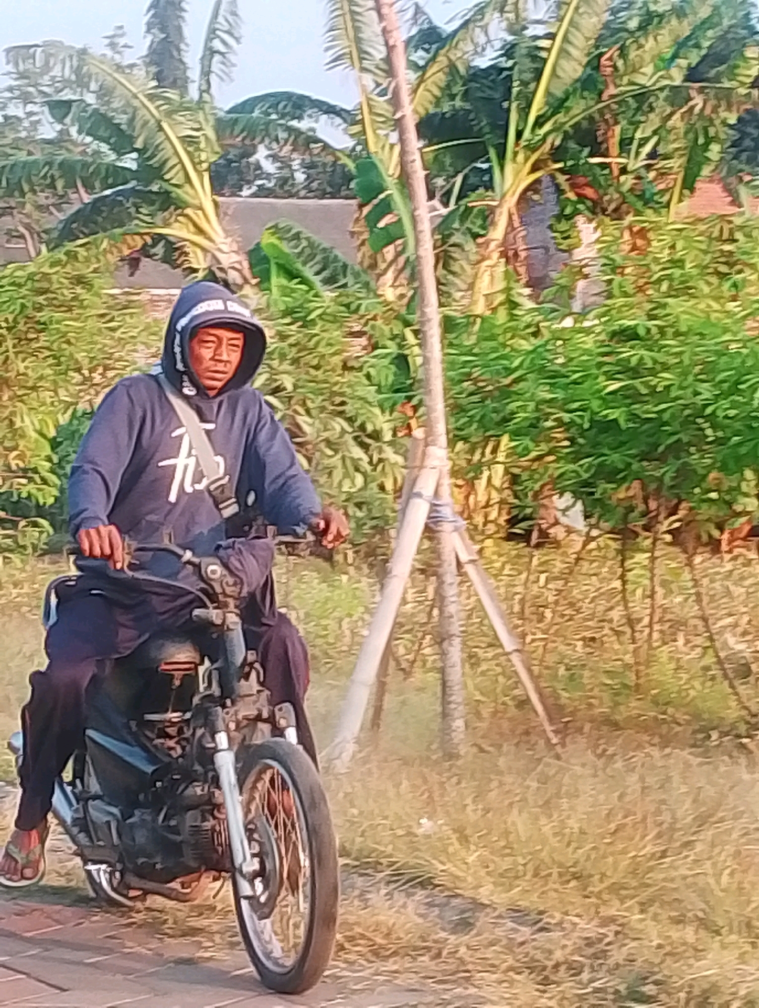 A man wearing a dark blue hooded sweatshirt and dark pants is riding a black motorcycle. The man is wearing a light-colored bag slung across his chest. He is wearing flip-flops on his feet. The motorcycle appears to be an older model with exposed engine parts. The background shows lush green foliage, including what appear to be banana trees, and a dirt path with dry grass. There is also a wooden post and what looks like bamboo supports in the background. The scene suggests an outdoor setting, possibly in a rural or semi-rural area.