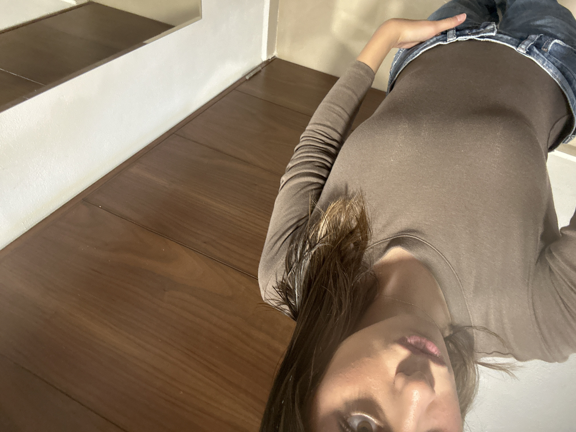 A close-up, high-angle shot shows a woman in a brown long-sleeved shirt and blue jeans. The background features a light-colored wall and a wooden floor or steps. The location is unspecified in terms of city and country.