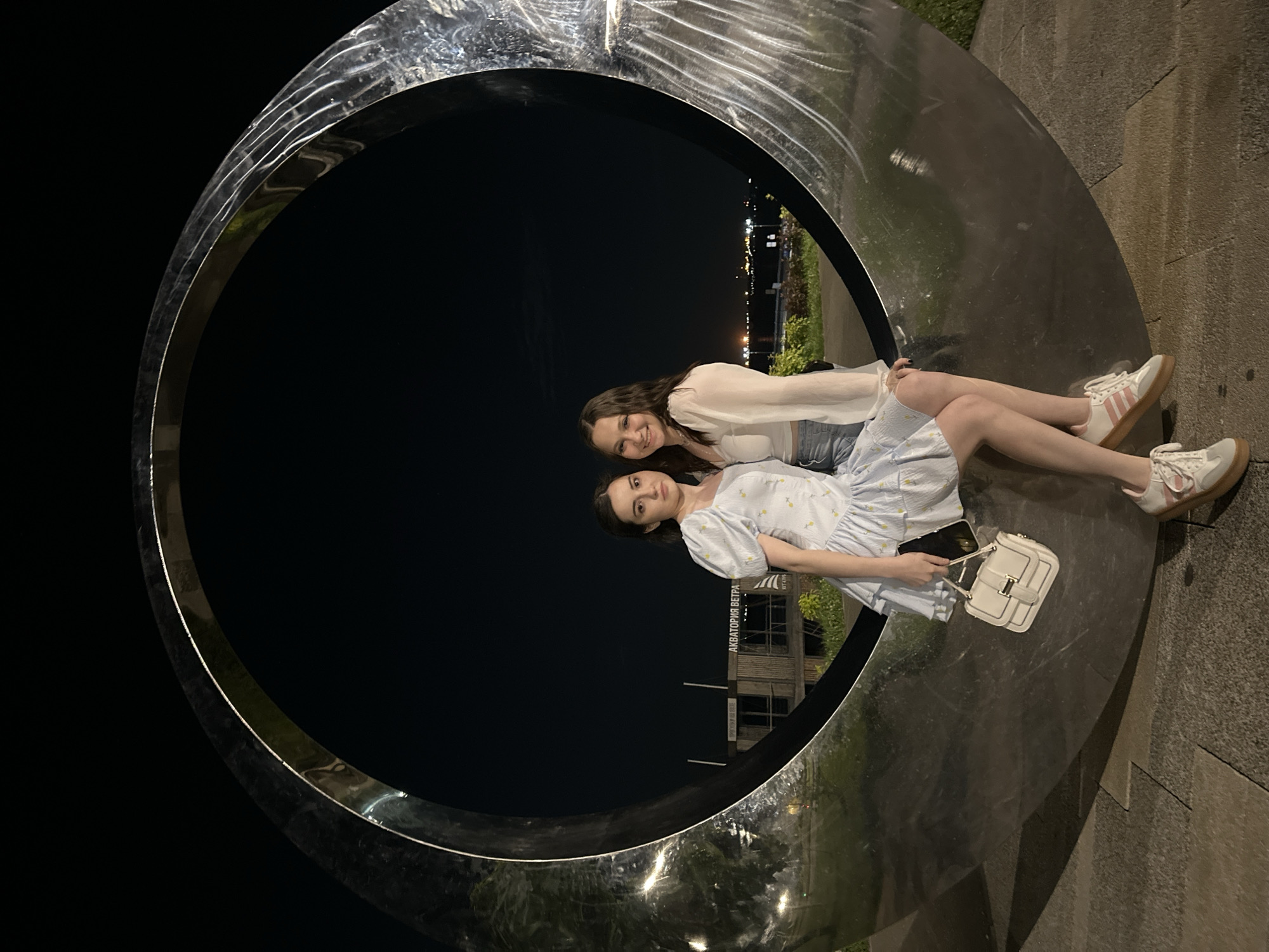 Two young women are seated within a large, circular, metallic sculpture at night. The sculpture appears to be an abstract art installation. The woman in front is wearing a light blue dress with a floral pattern and is holding a smartphone. She has dark hair and is looking towards the camera. Beside her, slightly behind, is another woman with long brown hair, wearing a white top and denim shorts. She is also smiling at the camera. Both women are wearing sneakers. The background shows a cityscape with lights and some greenery, suggesting an urban park or promenade. The time of day is clearly night, and the lighting is artificial. The overall mood appears relaxed and friendly. The text "АКВА TOPИЯ ВЕТРА" is visible on a building in the background. The scene is likely in Saratov, Russia, based on the location context provided.