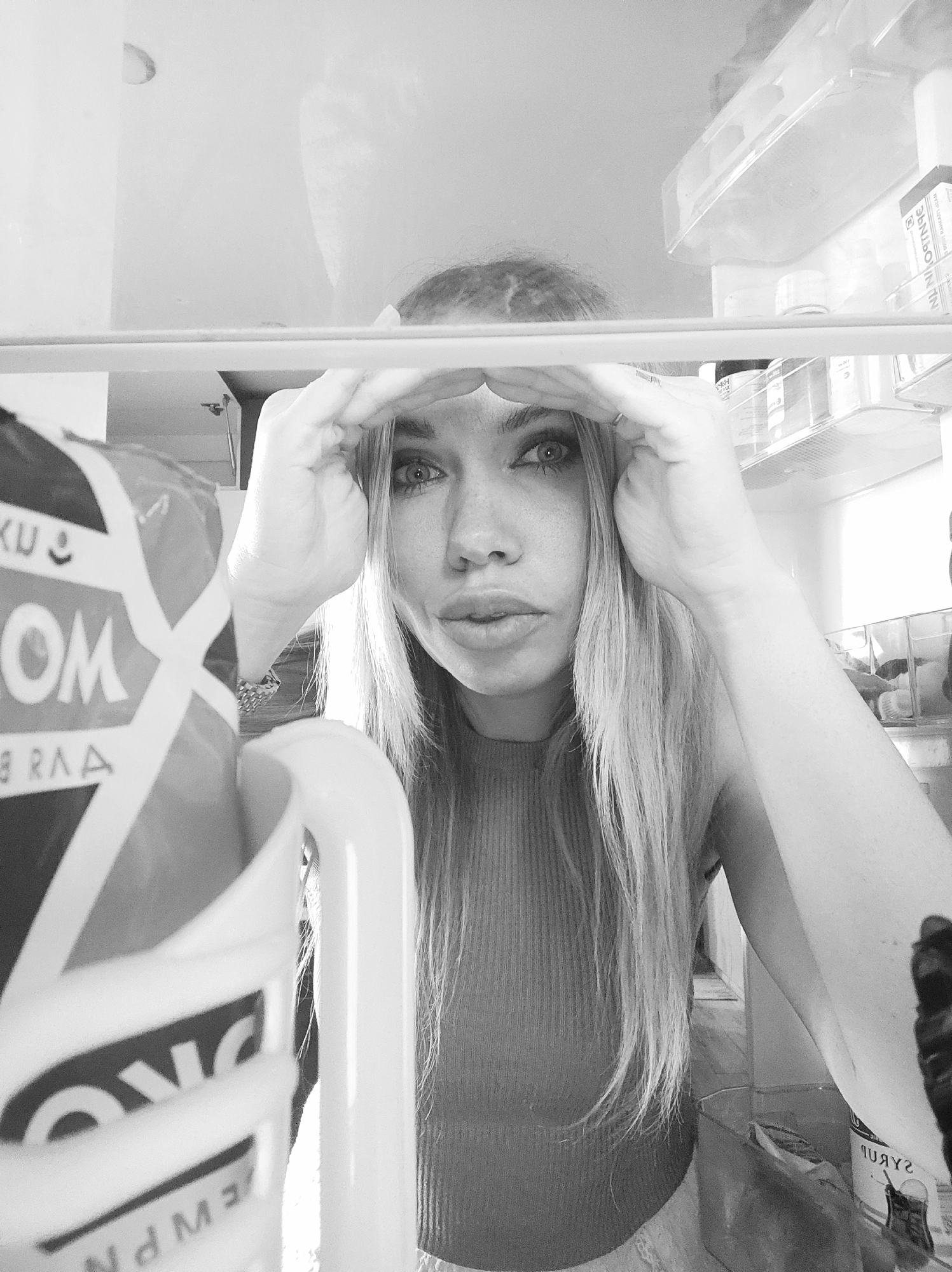 A grayscale, eye-level close-up shot from inside a refrigerator focuses on a fair-skinned woman with long, light hair. She is positioned centrally, gazing directly at the viewer with a slightly puckered expression. Her hands are raised, framing her forehead as if shielding her eyes.
Inside the refrigerator, various items are visible on shelves above and to the sides of the woman. These include packaged goods, jars, and containers. Part of the refrigerator door with the handle is in the foreground on the left side.
The location in Votkinsk, Russia is not known.
