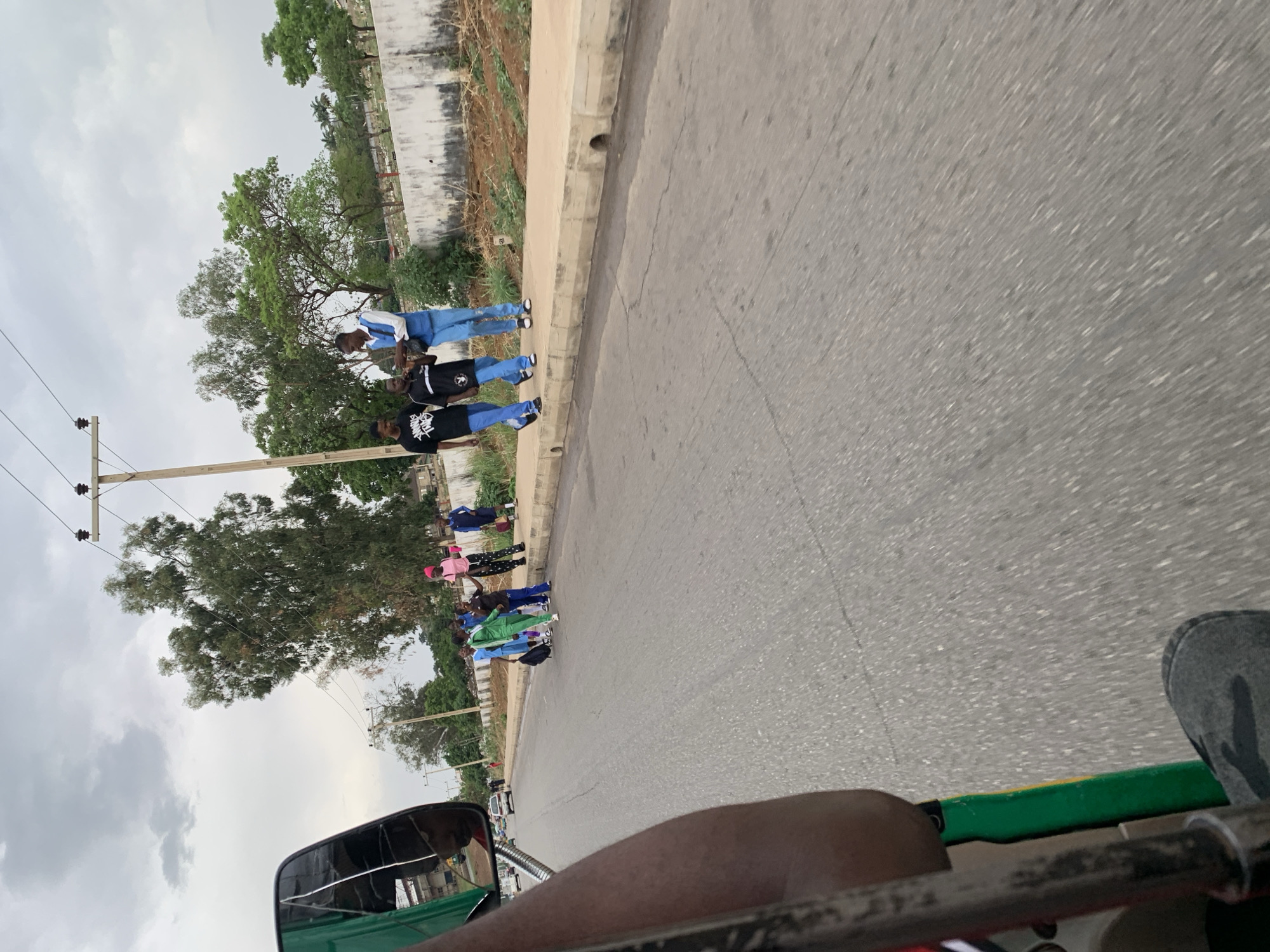 The image, oriented correctly, captures a roadside scene on an overcast day, likely in a semi-urban or suburban area in Nigeria. The photograph is taken from a low angle, apparently from inside a vehicle such as a tricycle (Keke Napep), with a dark-skinned arm and part of a green handlebar visible in the foreground.
In the midground, a paved road stretches diagonally from the bottom-left towards the top-right. Adjacent to the road, on the right, is a concrete sidewalk. A group of approximately ten individuals, primarily young adults or teenagers, are standing along this sidewalk. They are dressed casually; notable attire includes several people in blue jeans with various tops, including one in a white long-sleeved shirt, and two individuals in black t-shirts, one of which has a discernible white logo that resembles "STUSSY" branding. Further down the line, other individuals are seen in bright casual wear, including tracksuits in shades of green and blue, and one person in a pink top and patterned pants. They appear to be waiting or congregating by the roadside.
Beyond the sidewalk, there's a strip of grass and a low, weathered concrete wall. Further into the background, mature trees with dense green foliage are visible, along with power lines and utility poles. The sky is largely covered with grey clouds, indicating an overcast or cloudy day. No specific activity beyond standing is clearly depicted, and no strong emotions are discernible from the individuals. The overall setting is consistent with a public street or thoroughfare near a place like a stadium.
