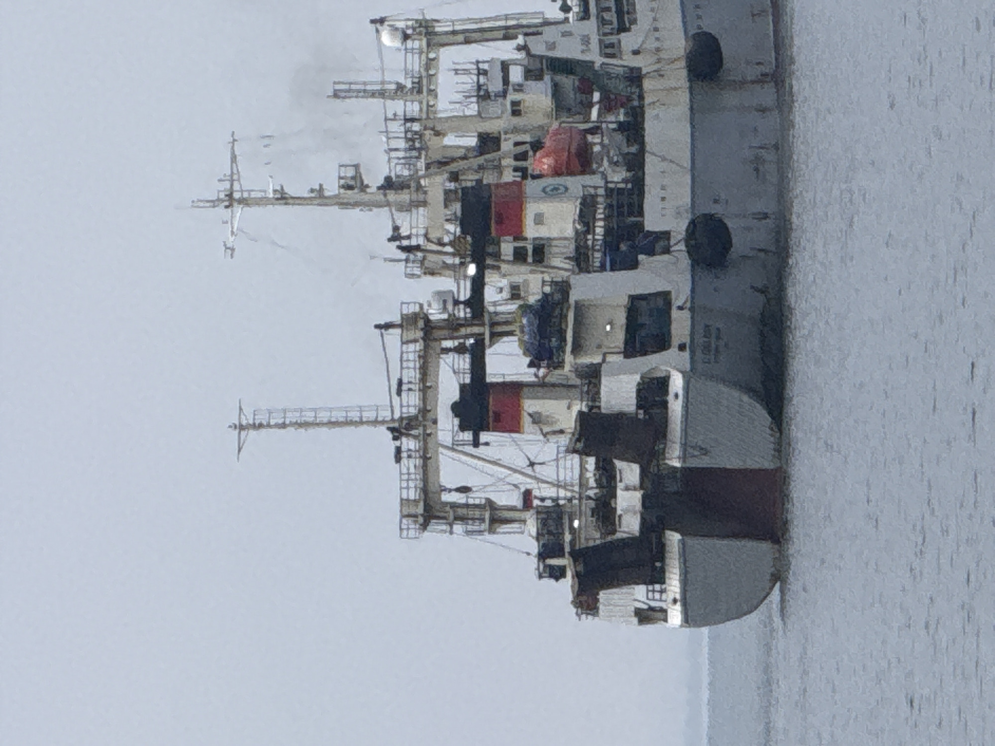 An industrial vessel, likely a fishing trawler or research ship, is stationary in calm, grey water, possibly in a harbor or bay. The image is taken on an overcast day, indicated by the muted light and lack of strong shadows, with the sky appearing uniformly grey.
The vessel is large and complex, featuring multiple decks, a prominent superstructure, several cranes, masts, and antennas. Smoke or steam is rising from a stack near the center of the ship, suggesting it is operational or has recently been in use. The hull is predominantly white and light grey, with a dark red stripe marking the waterline, and some sections of the superstructure are dark grey or black. Red accents are visible on some equipment covers or panels. Black fenders are hanging along the side of the hull. Various pieces of equipment, including what appears to be radar domes and davits, are visible on the upper decks.
No people are visible in the image. The water is smooth and reflects the overcast sky, indicating calm conditions. The overall atmosphere is quiet and industrial.
Visible text on the hull, near the stern, appears to be in Cyrillic script, faintly showing "РВ БОРИС МИ" and "Б-0062", likely indicating the ship's name ("Boris M." or similar) and a registration number. Other small, indistinct text is present on the superstructure.