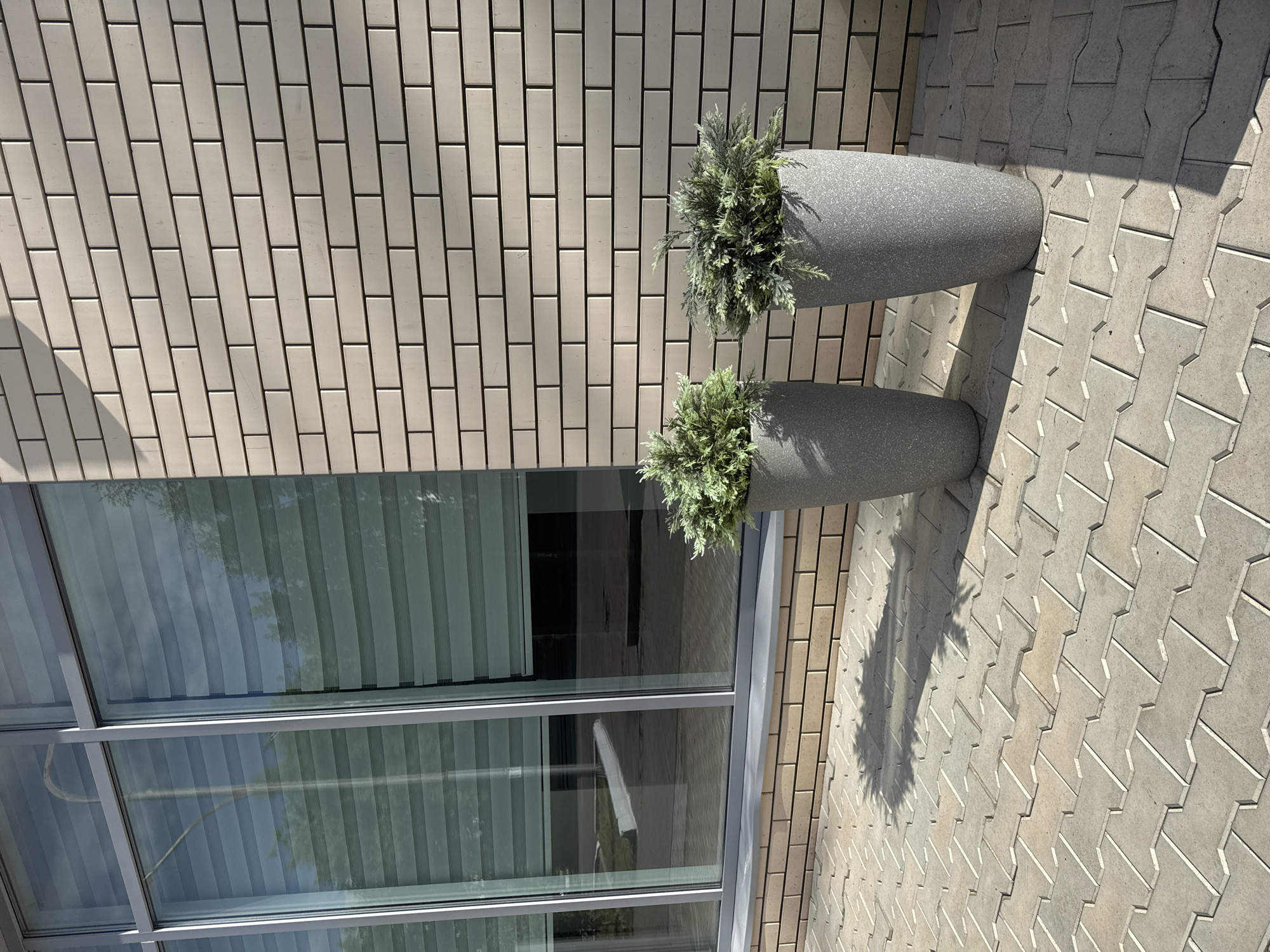 Two gray, textured, conical planters filled with low-lying green shrubbery stand on a paved walkway in front of a building. The building features a wall of light beige brick and a large window with horizontal blinds. The walkway is made of interlocking concrete pavers. A shadow of a tree is cast on the walkway. The scene appears to be an outdoor area of a commercial or public building.