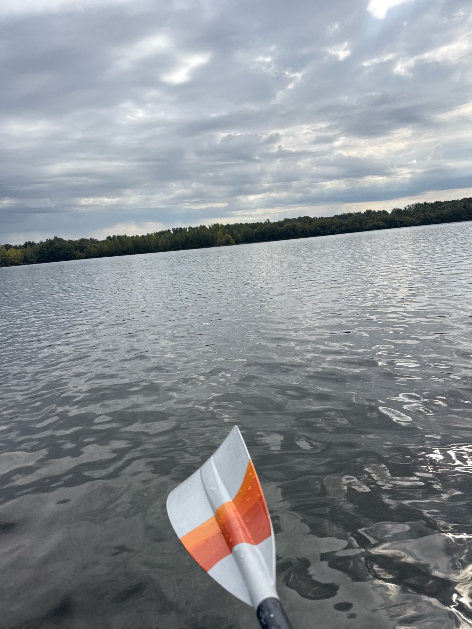 An overcast sky with scattered clouds hangs above a calm body of water, likely a lake or wide river. The water's surface is rippled, reflecting the muted light. In the foreground, the tip of a paddle with orange and white markings is visible, suggesting a person is in a watercraft. A dense line of trees forms a dark horizon in the distance. The image does not provide any specific landmarks or clues to pinpoint its location within the city of Ufa, Russia.