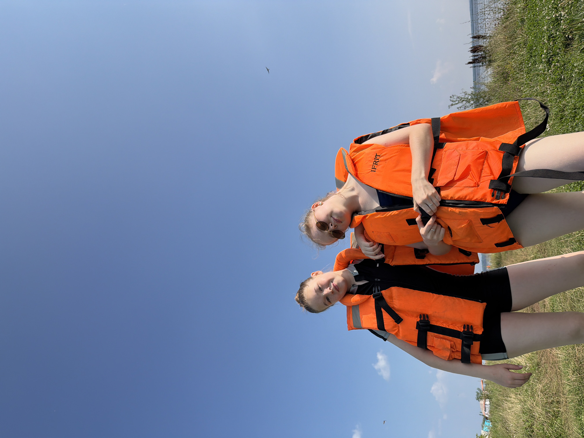 Two young females, dressed in black swimsuits and bright orange life vests, stand outdoors with a clear blue sky and a body of water in the background. The female on the left is leaning on the other, with her head tilted slightly back and a faint smile. The female on the right is looking towards the camera with sunglasses on and a relaxed posture. Both are wearing orange life vests with black straps and buckles. The location is identified as Krasny Bor, Russia, suggested by the presence of a grassy embankment leading to a body of water, characteristic of the region's natural landscape. The scene appears to be a recreational outing, possibly near a beach or riverbank. The overall lighting indicates it is daytime.